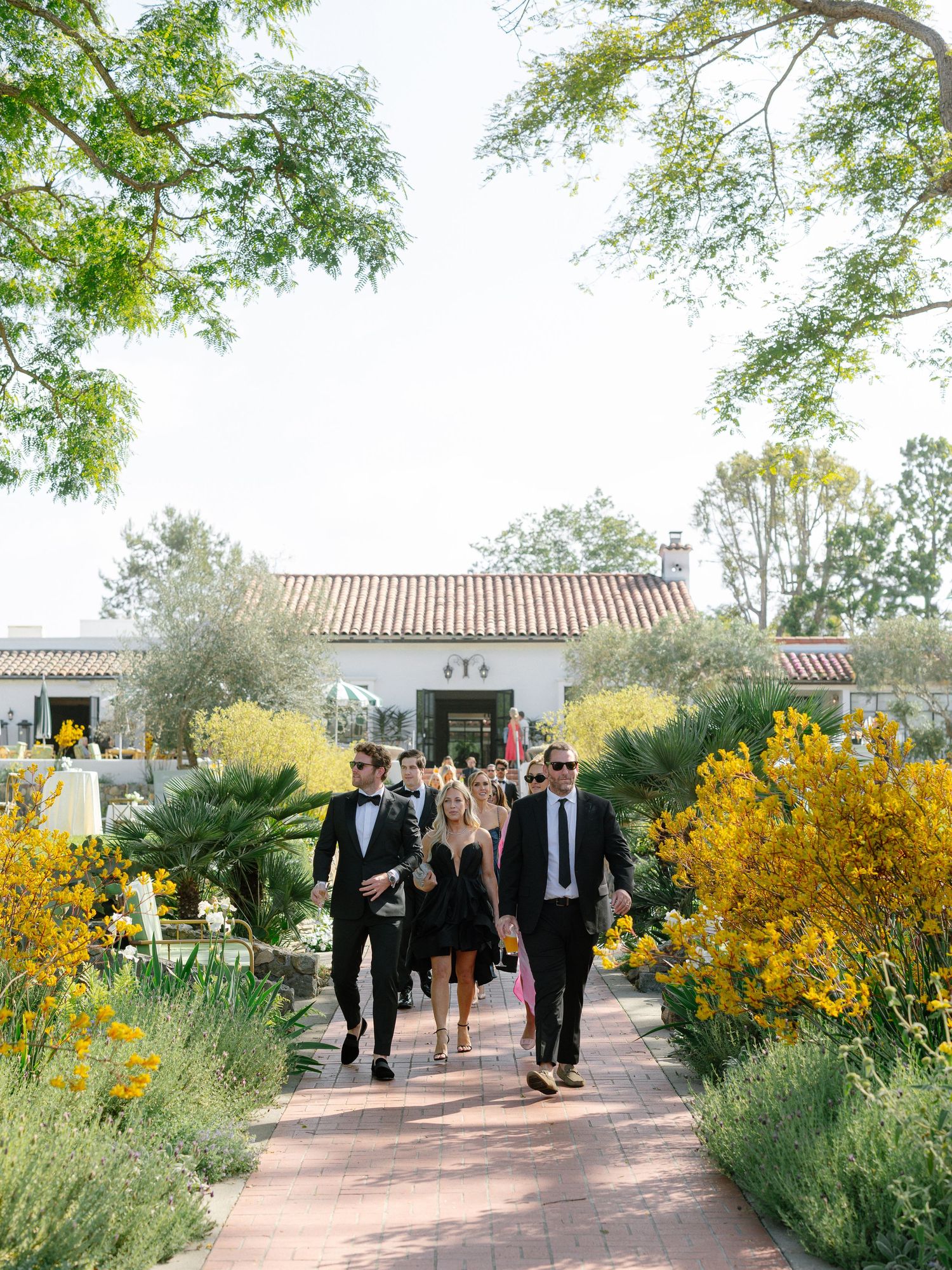 Wedding guests in formal attire walking along a brick path through a lush garden venue.