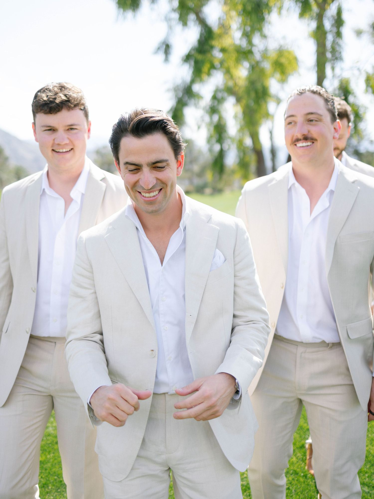 Three groomsmen in matching cream linen suits smile and walk together outdoors on a sunny wedding day.