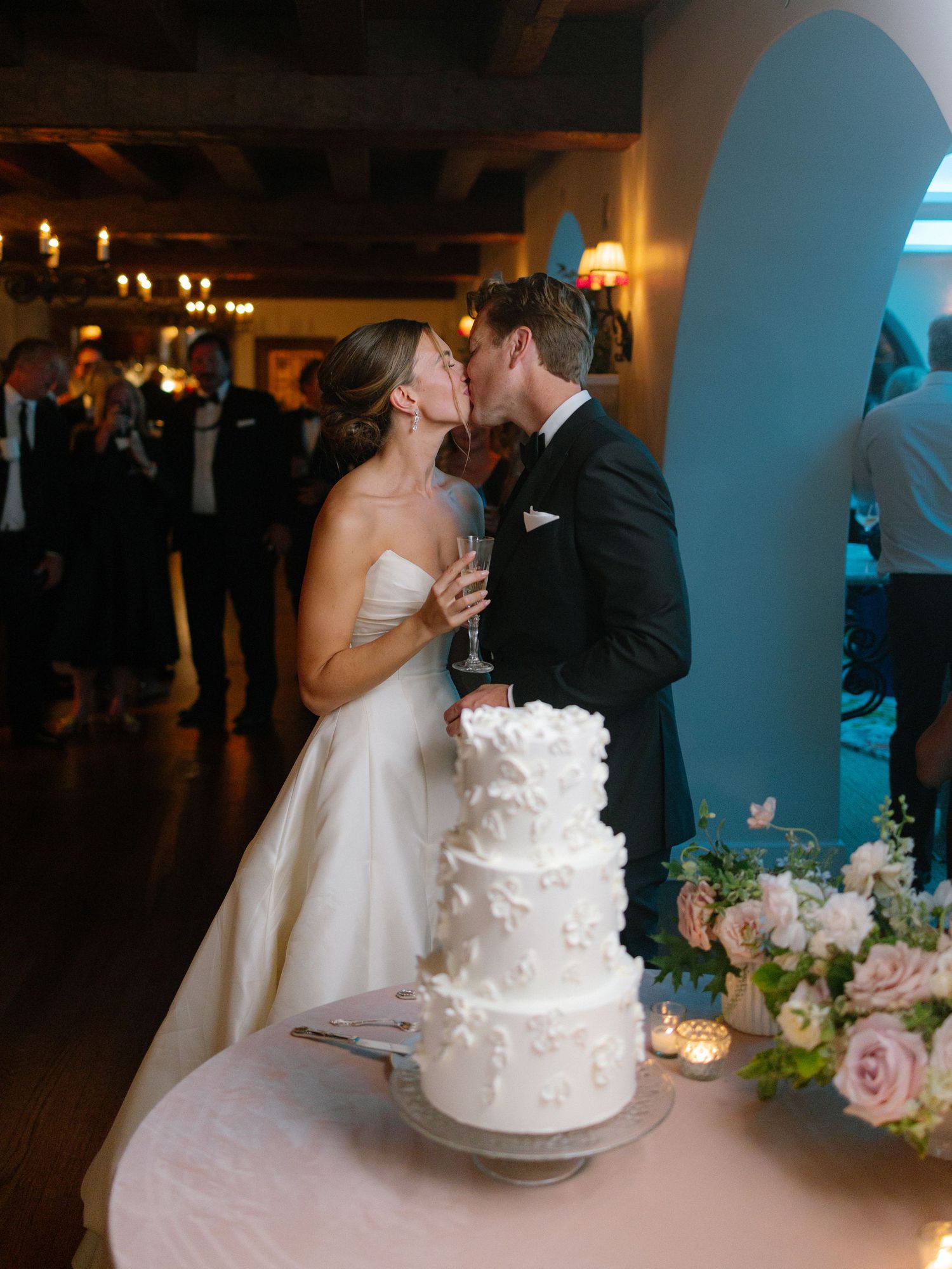 Newlyweds kiss over three-tier white floral wedding cake during romantic indoor reception celebration.