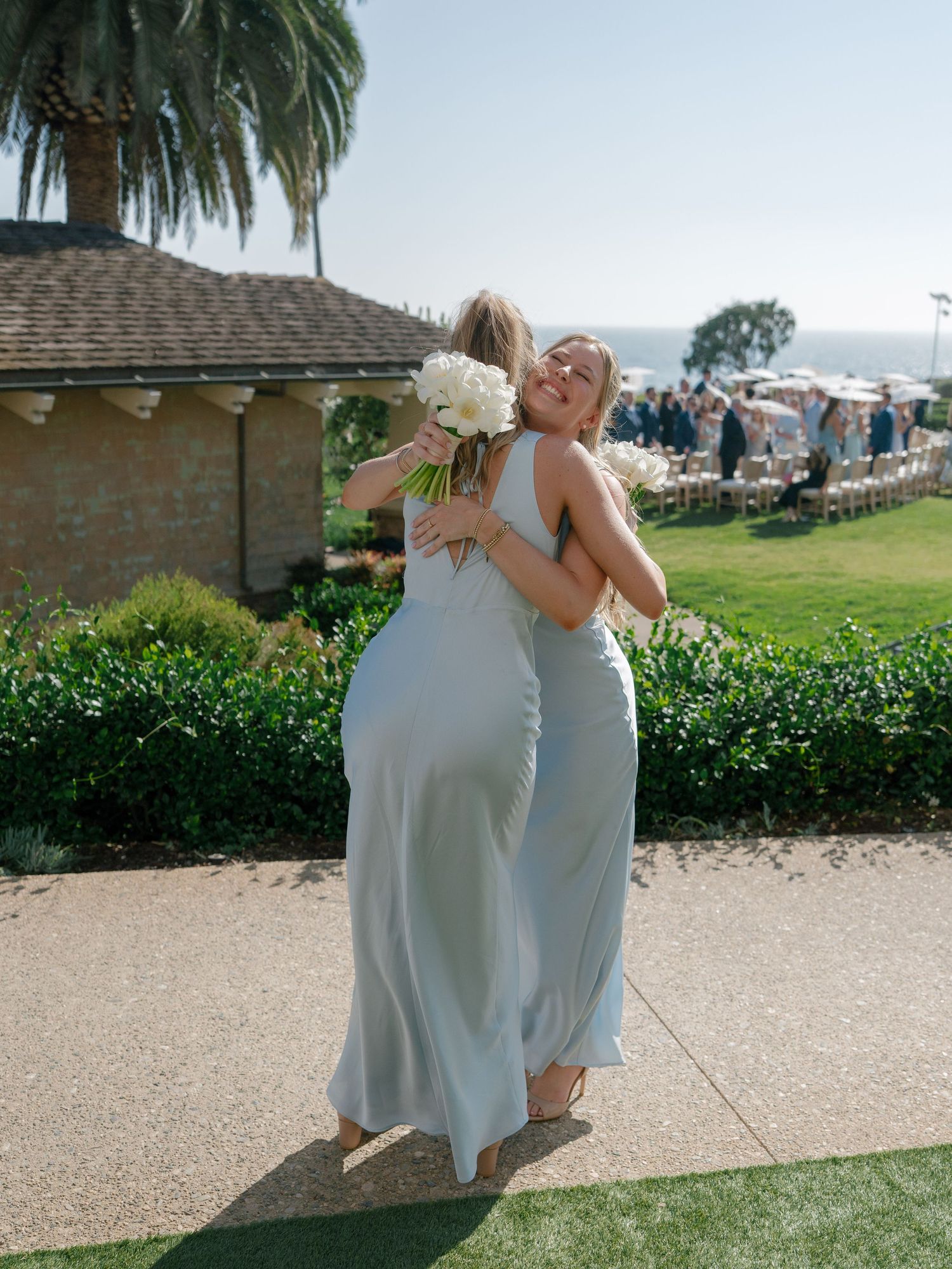 Two bridesmaids in sage green gowns embrace outdoors at a coastal wedding venue with ocean views.