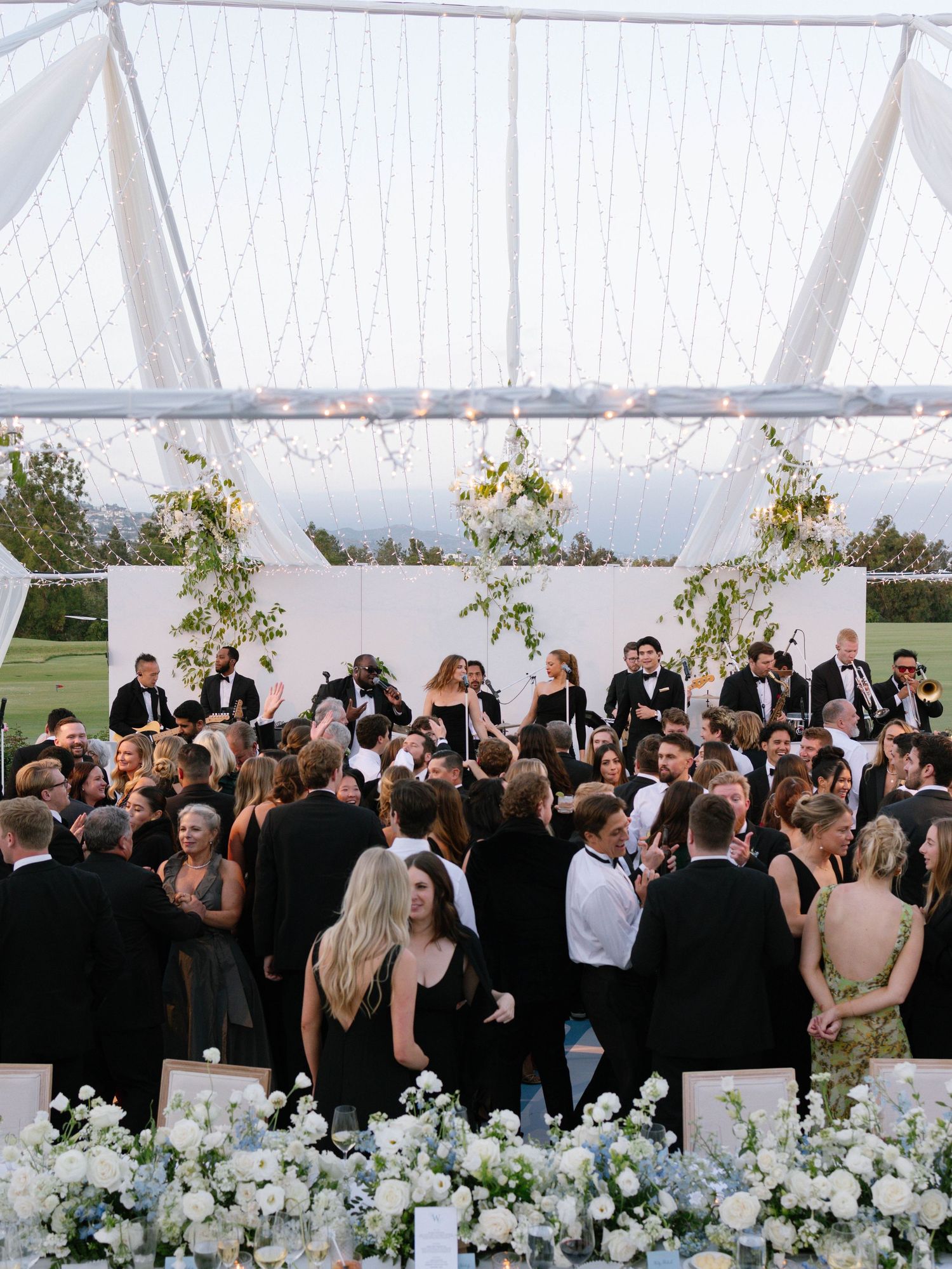 Aerial view of elegant wedding reception under clear tent with guests dancing and white floral decor.