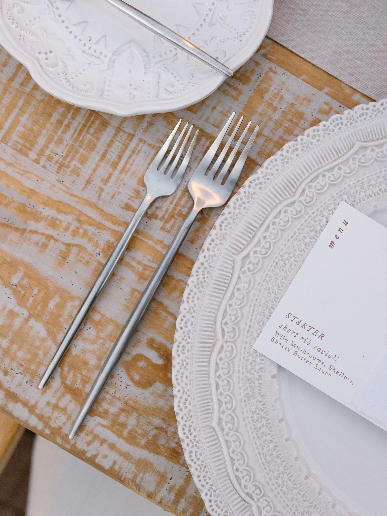 Close-up of silver forks beside a white lace-edged charger plate and printed menu card on a rustic wood table.
