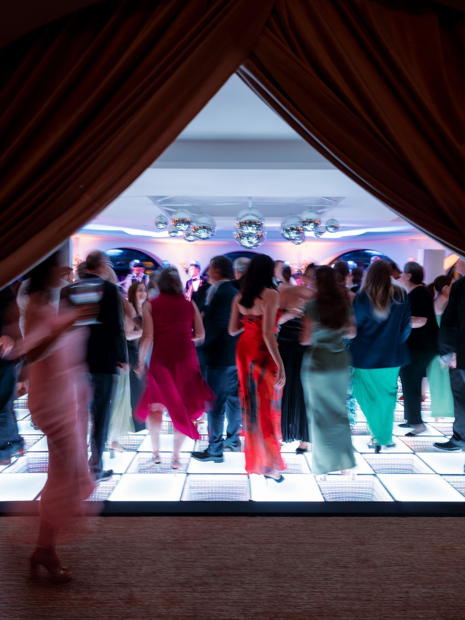 Wedding reception guests dancing on illuminated white dance floor viewed through draped curtain entrance.