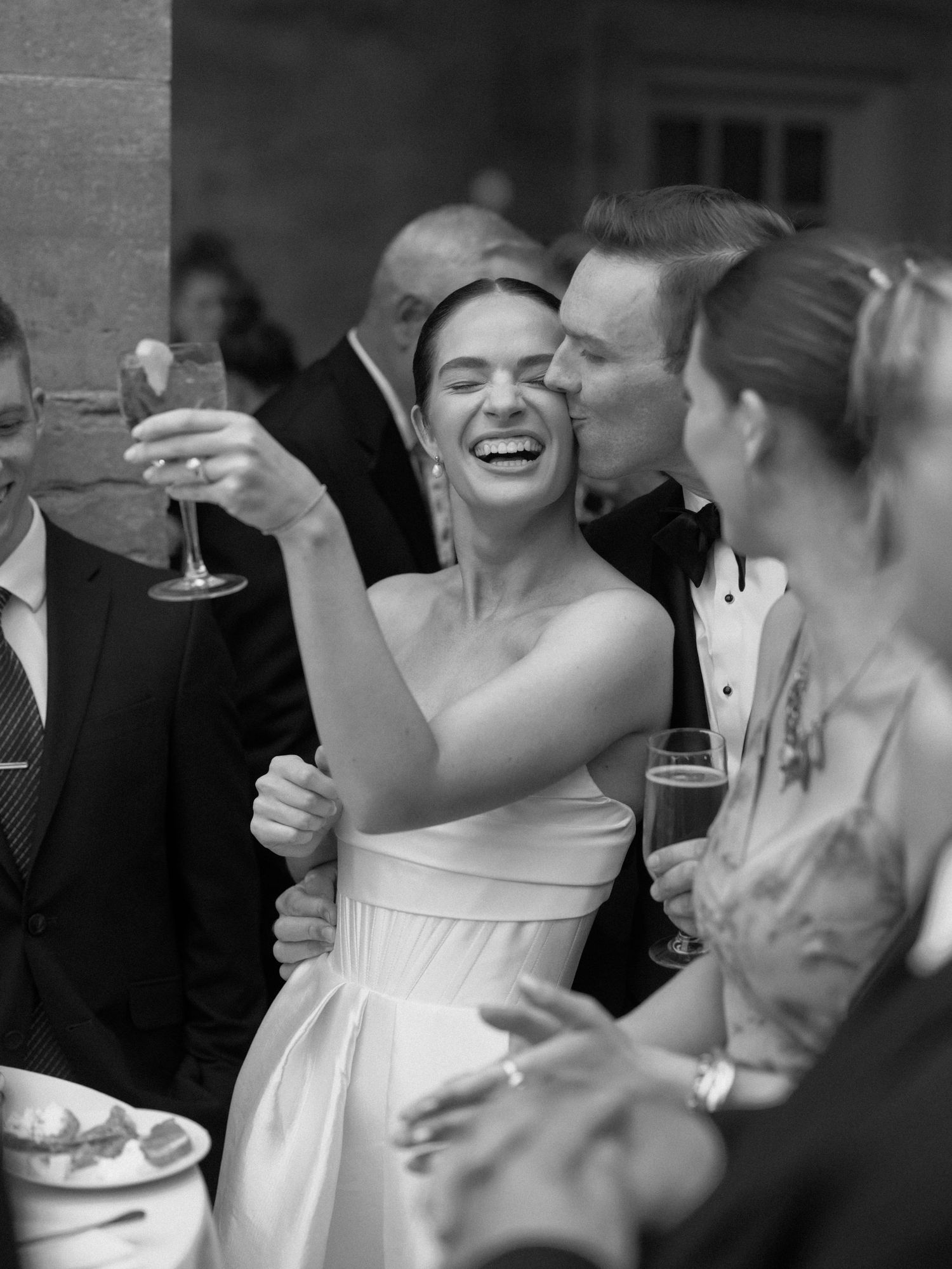 Bride laughing joyfully while holding champagne as groom kisses her cheek at wedding reception.