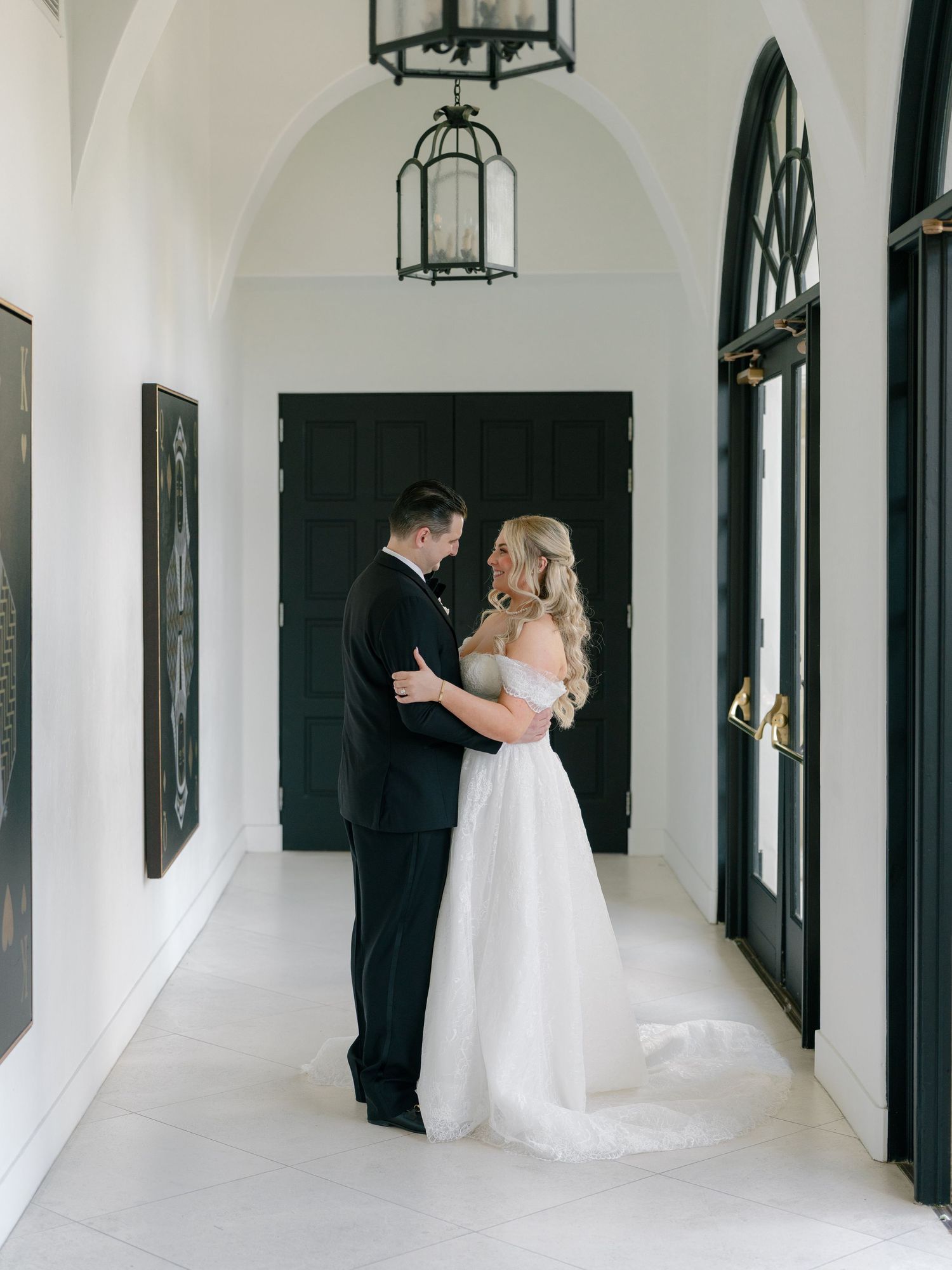 Bride and groom share tender first look moment in elegant white hallway with black doors.