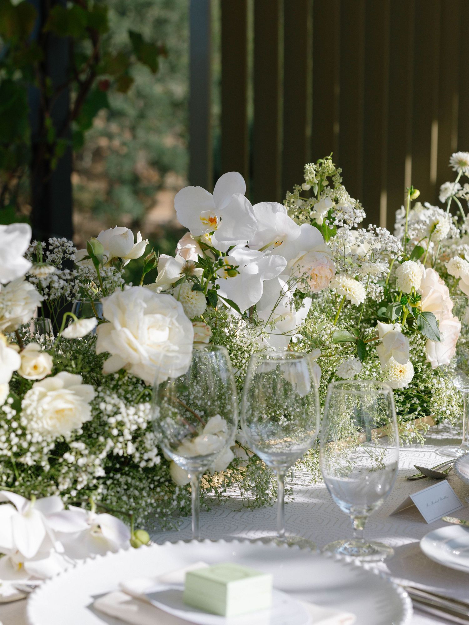 Lush white wedding tablescape featuring orchids, roses, and baby's breath arrangements on a white linen table setting.