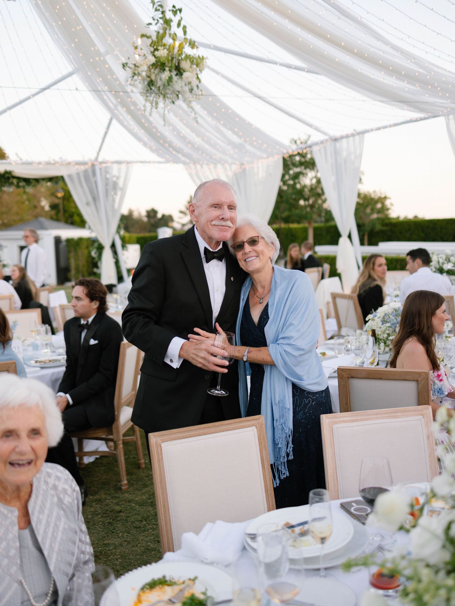 Elegantly dressed couple poses and smiles together at an outdoor tented wedding reception with white draped decor.