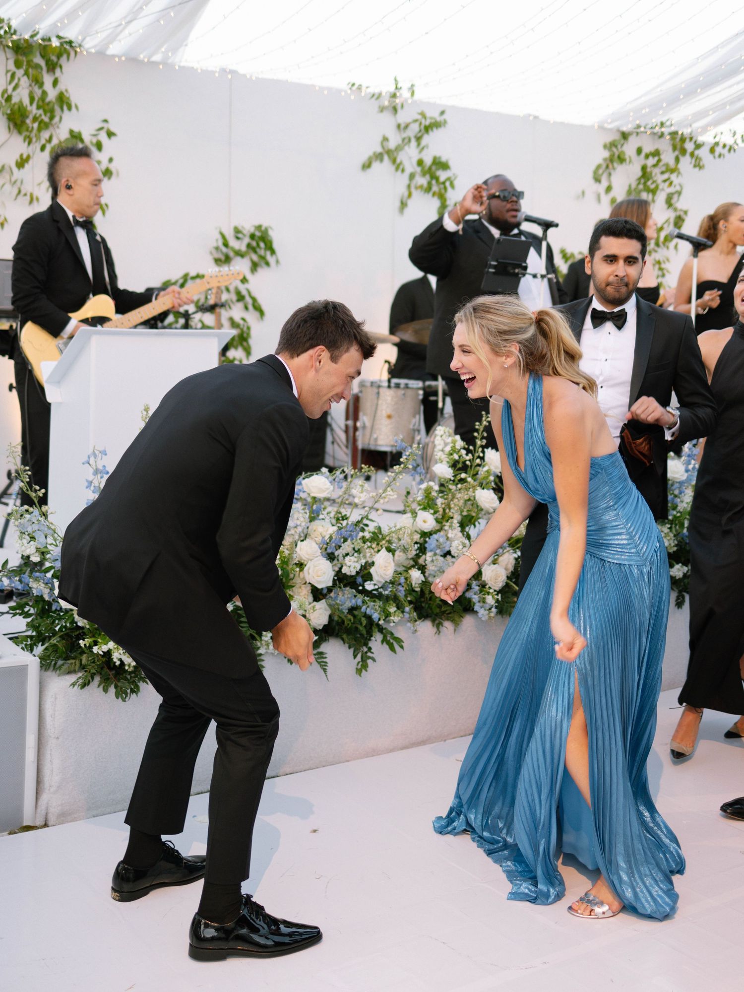 Wedding guests in blue pleated gown and tuxedo laughing together near floral arrangements and live band.