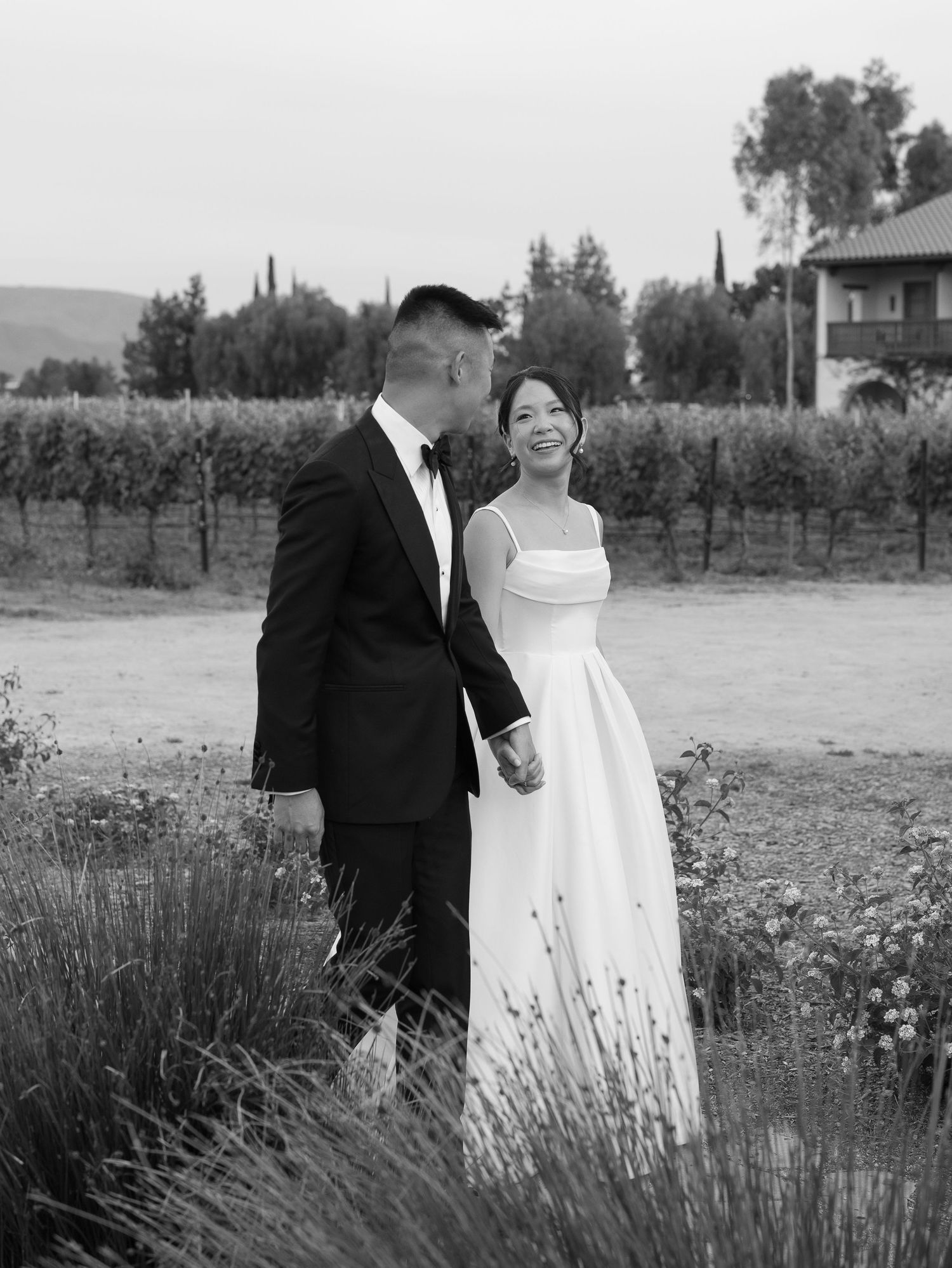 Black and white photo of smiling bride and groom holding hands while walking through a vineyard at dusk.