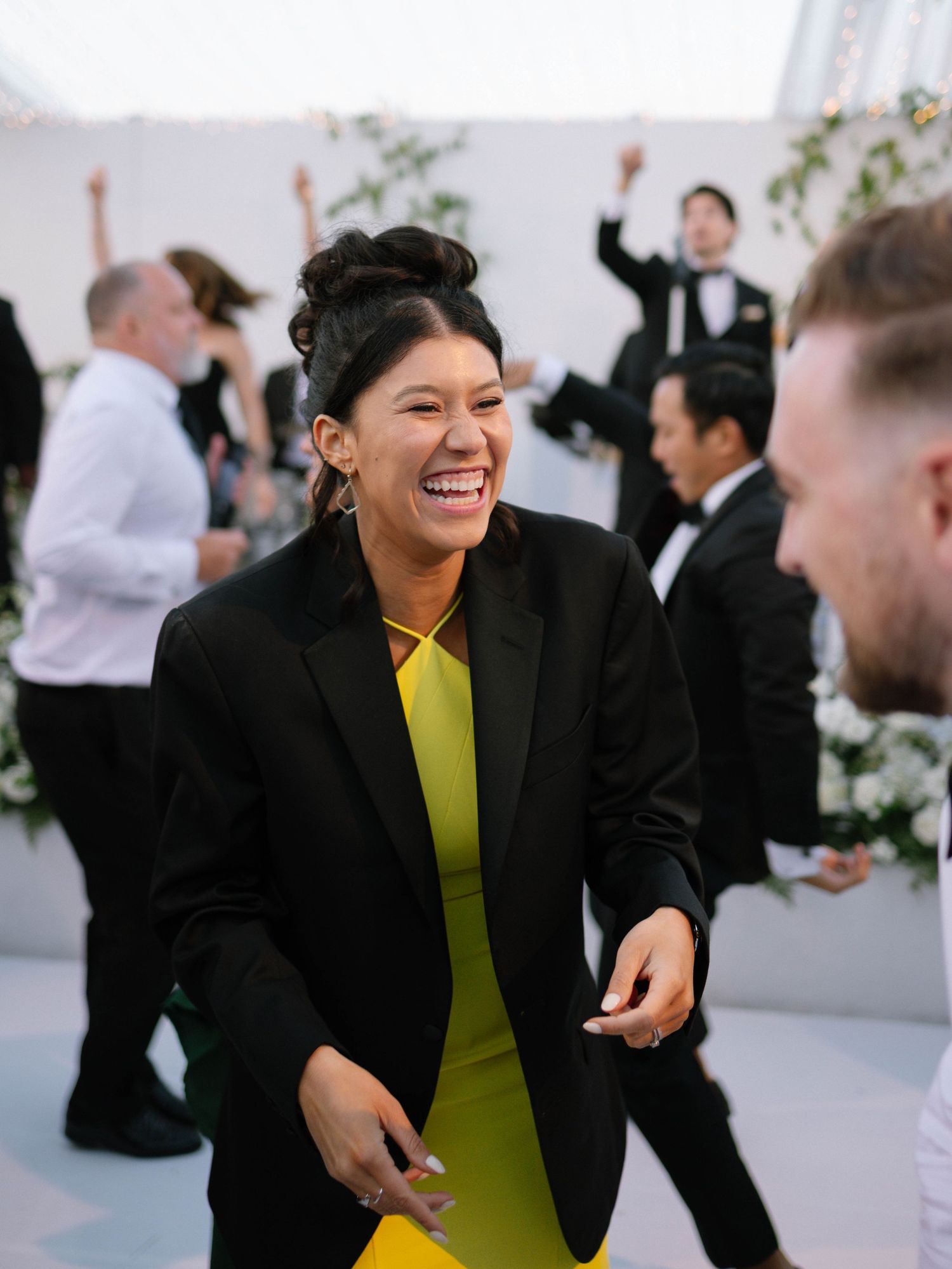 Wedding guest laughing in yellow dress and black blazer on dance floor during lively reception.