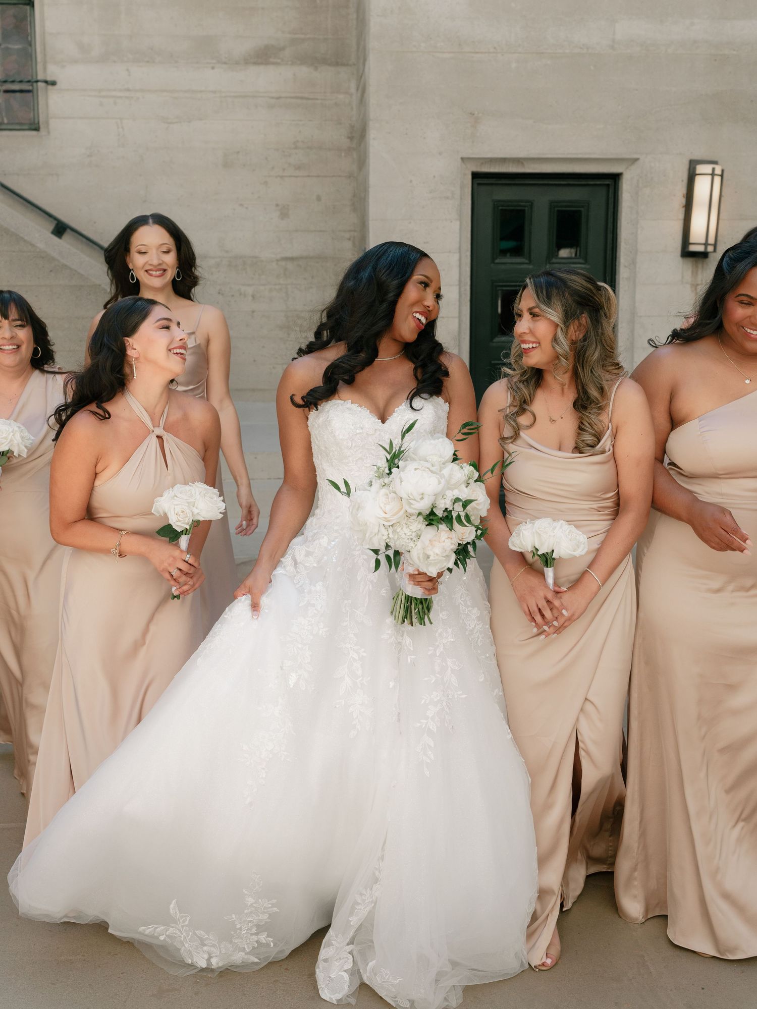 Bride in strapless lace gown laughs with bridesmaids in neutral satin dresses outdoors.