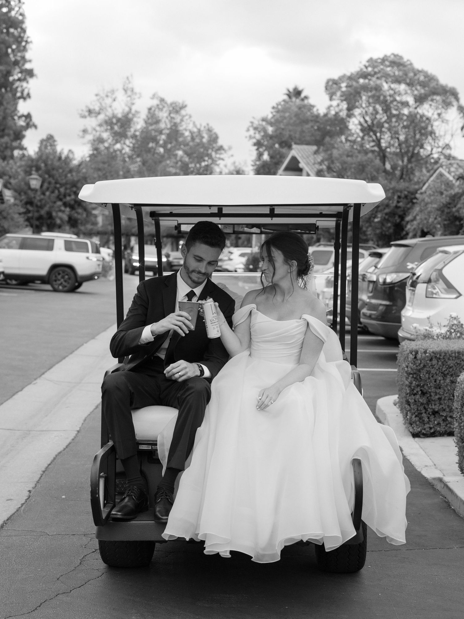Black and white photo of bride and groom sharing a toast while riding a golf cart on their wedding day.