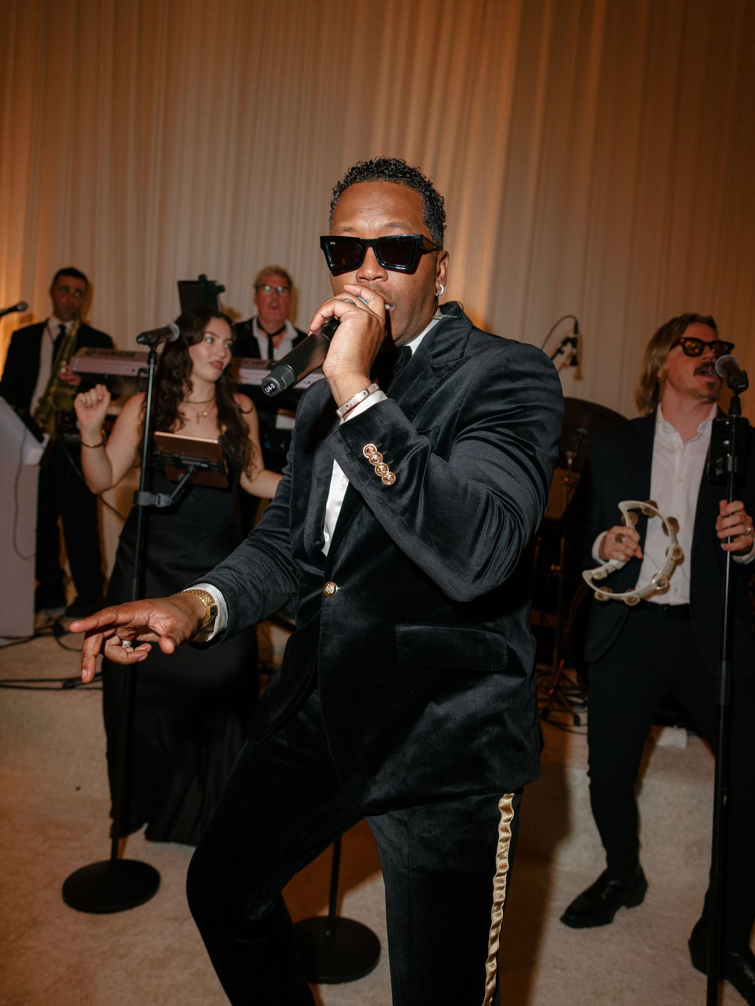 Man in black velvet suit and sunglasses performing with microphone at elegant wedding reception.