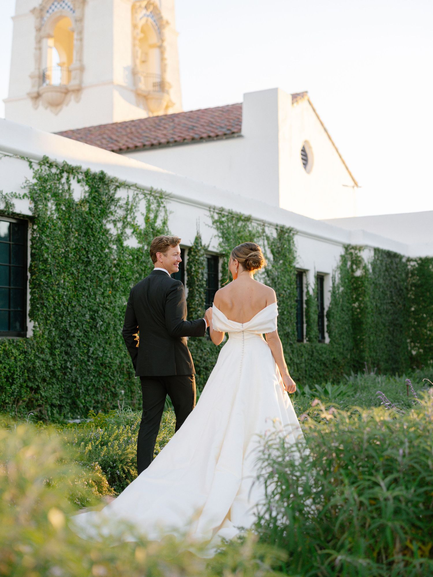Bride in off-shoulder gown with cathedral train walks with groom past ivy-covered chapel at sunset.
