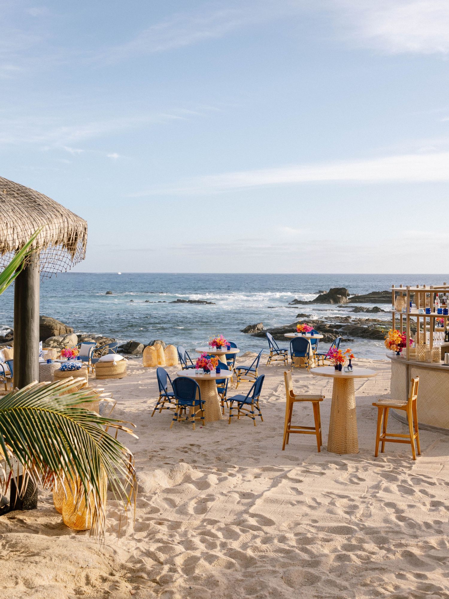 Colorful beachfront wedding reception setup with blue chairs, floral centerpieces, and ocean backdrop on sandy shore.