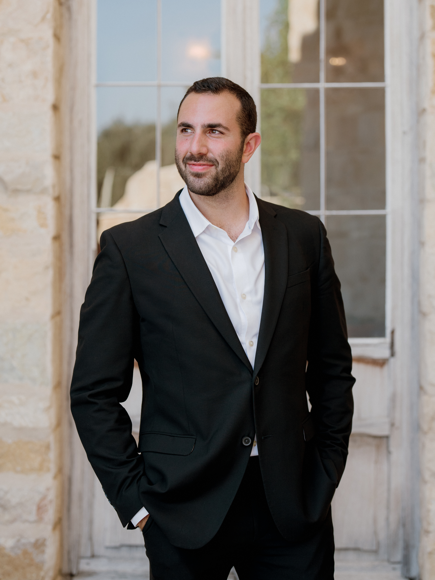 A well-dressed man in a black suit and white shirt stands confidently near elegant French doors outdoors.