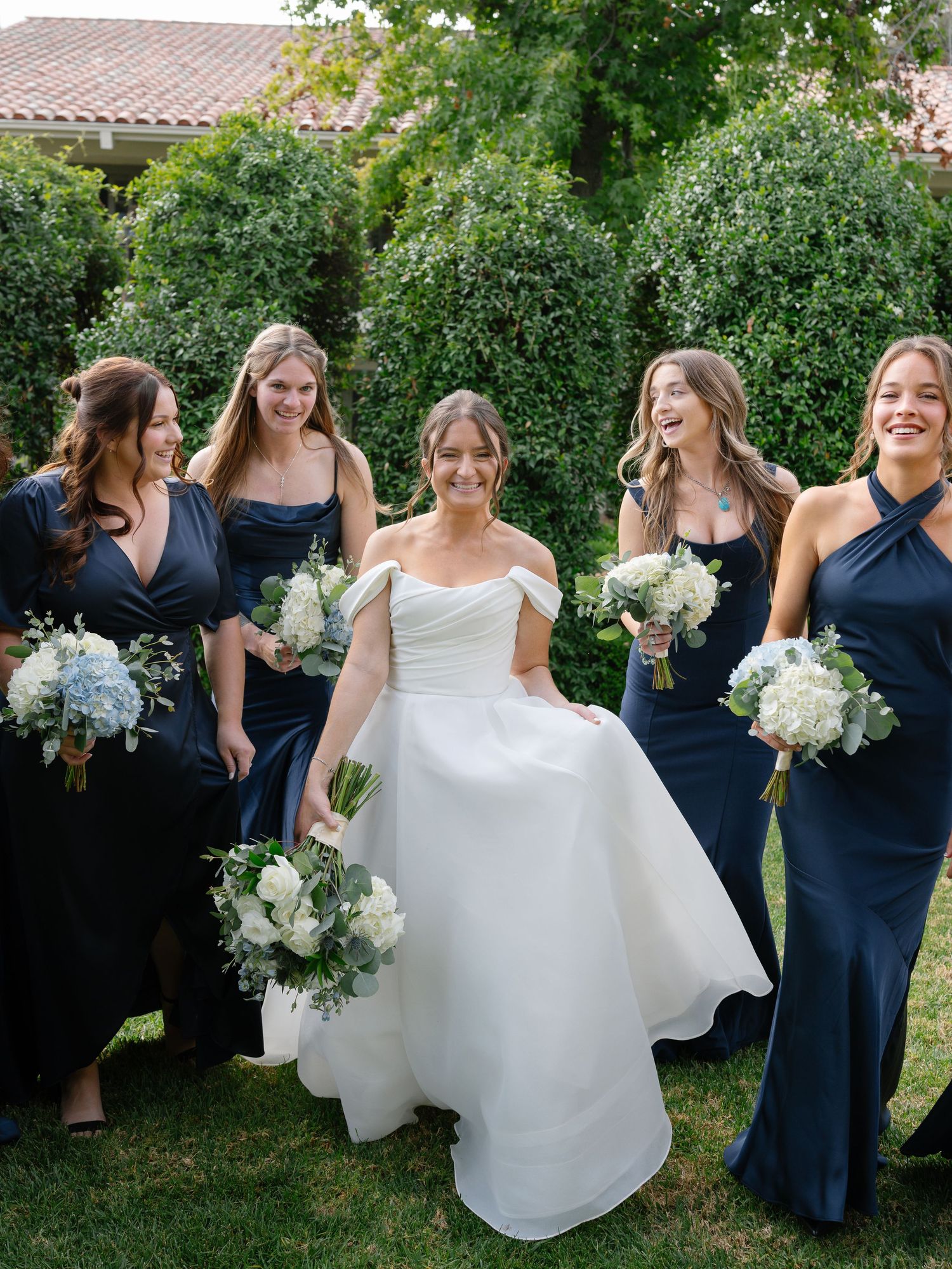 Bride in off-shoulder gown laughing with bridesmaids in navy dresses holding white hydrangea bouquets outdoors.