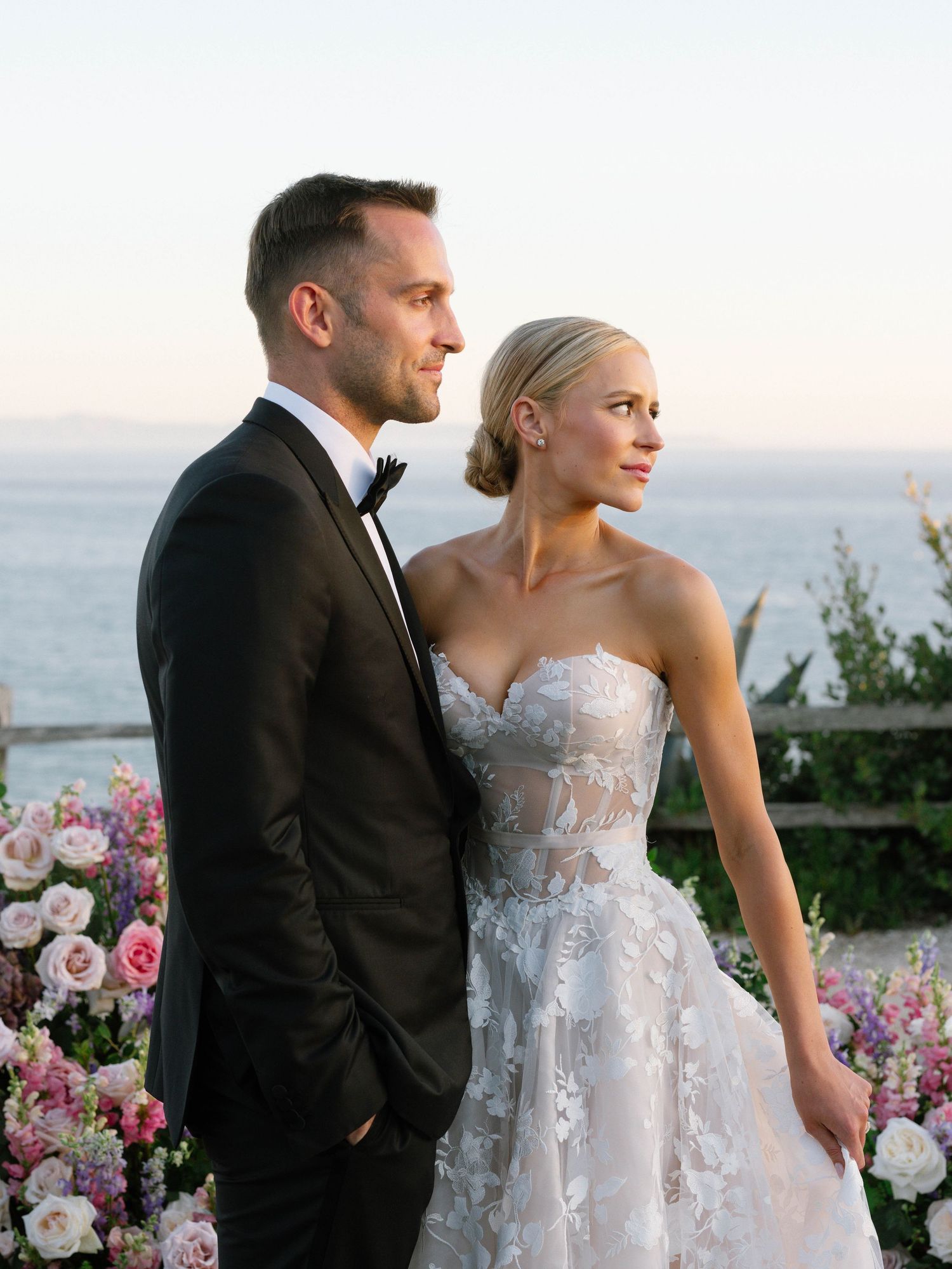 Bride and groom stand elegantly at sunset by the ocean, she wears a floral applique strapless gown.