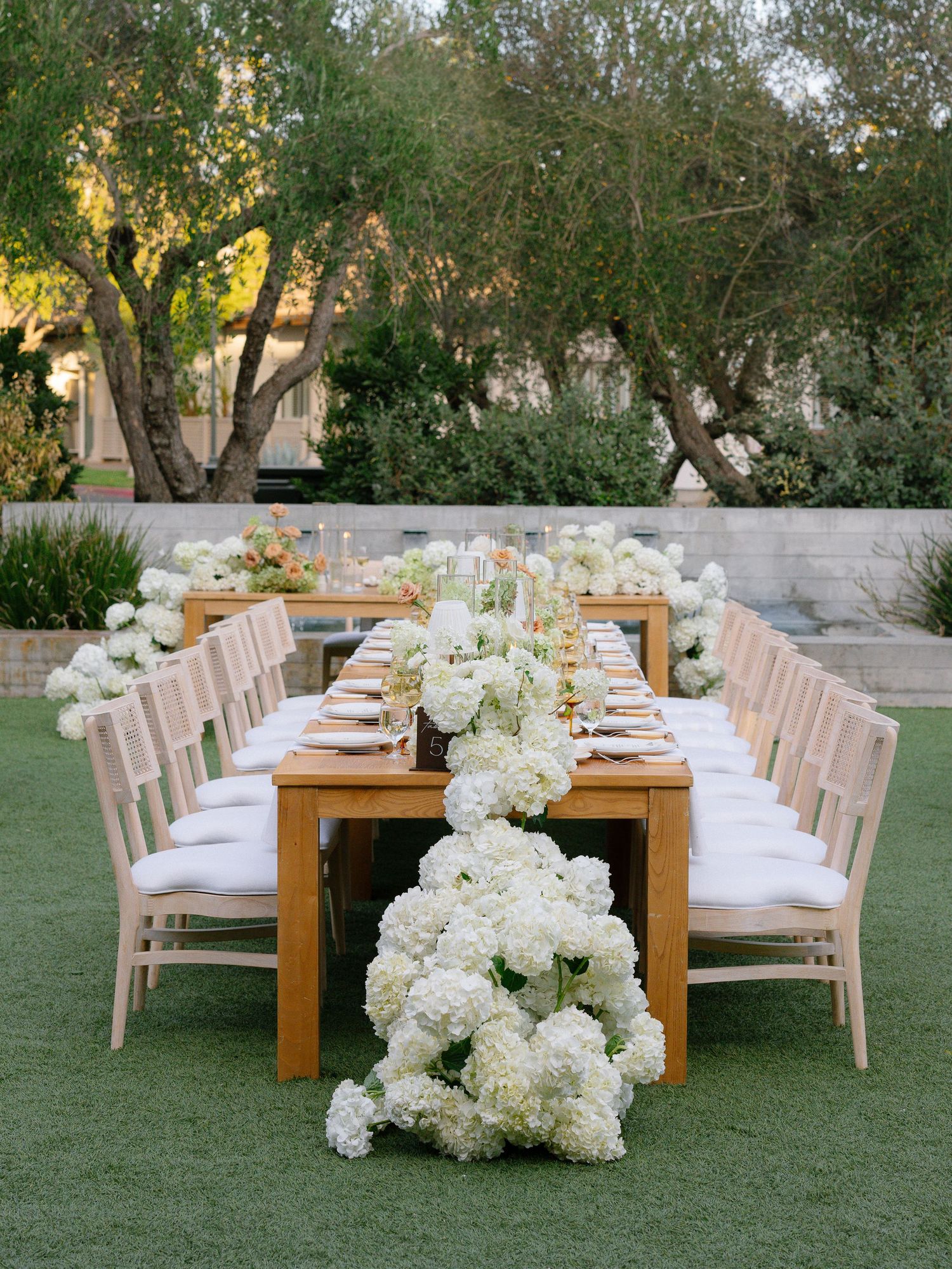 Elegant outdoor reception table with white hydrangea floral runner on green lawn.