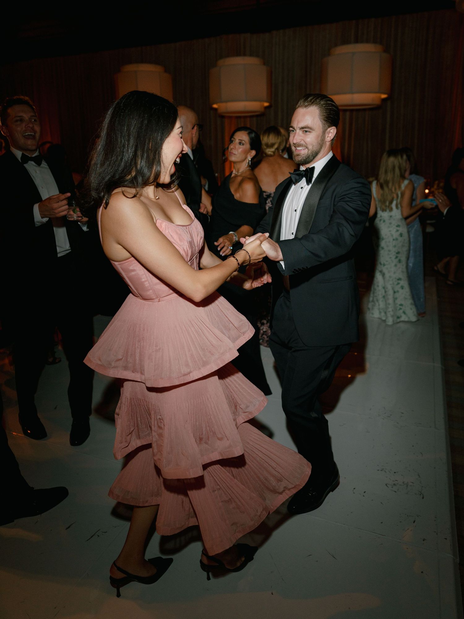 Wedding guests dancing at reception, woman in pink tiered dress dancing with man in black tuxedo.