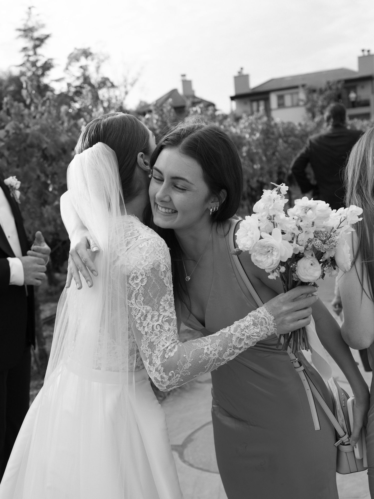 Guest warmly embraces bride in lace wedding gown at outdoor vineyard wedding ceremony.