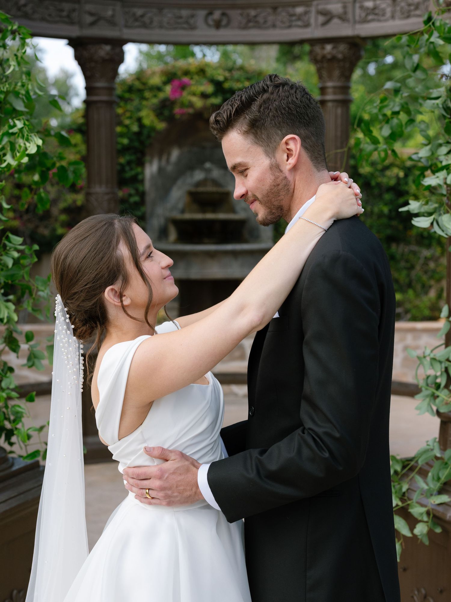 Bride and groom sharing romantic embrace under ornate garden gazebo with lush greenery backdrop.