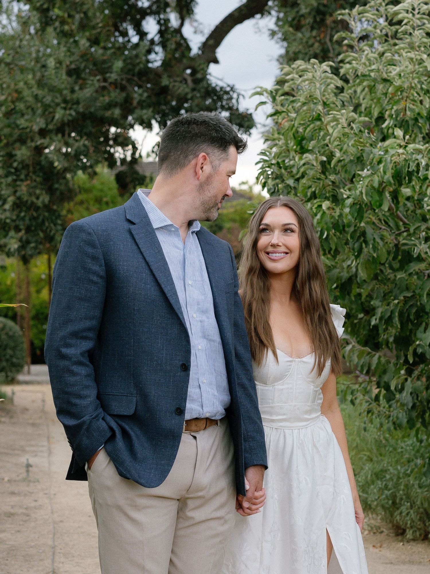 Smiling couple holds hands while strolling along a garden path surrounded by lush trees during engagement photos.