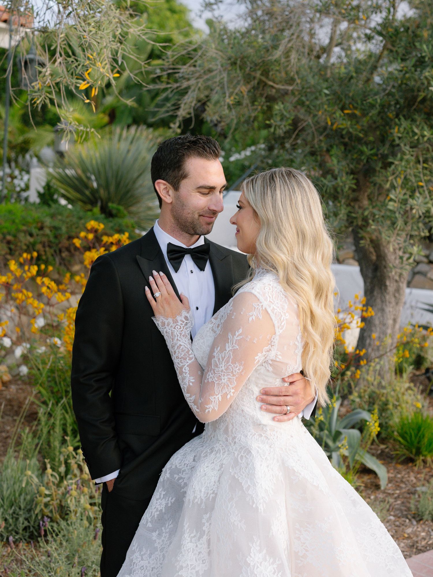 Bride in lace long-sleeve gown and groom in tuxedo share tender moment in garden setting.