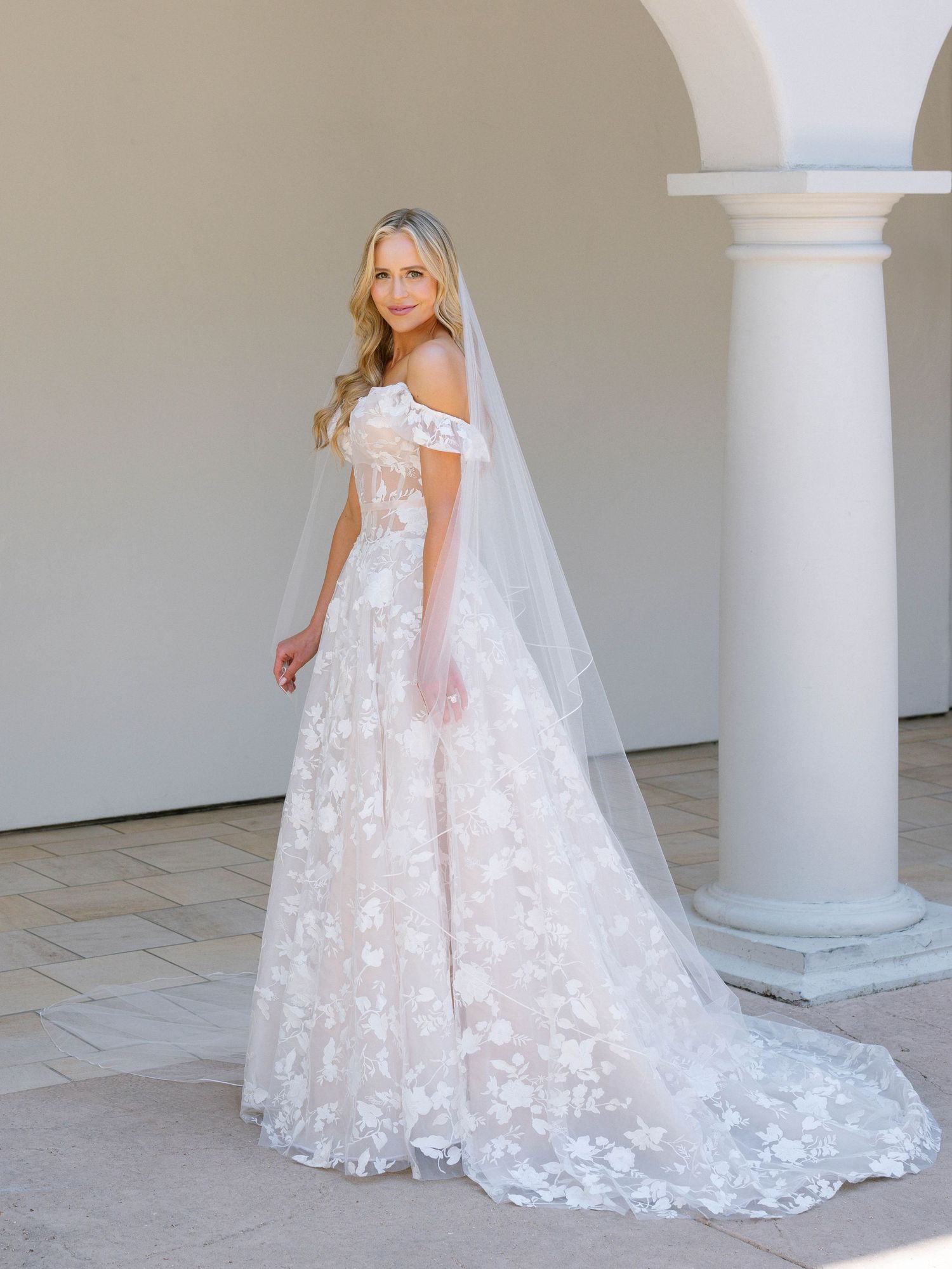 Blonde bride posing in off-shoulder floral lace ball gown with cathedral veil near stone columns.