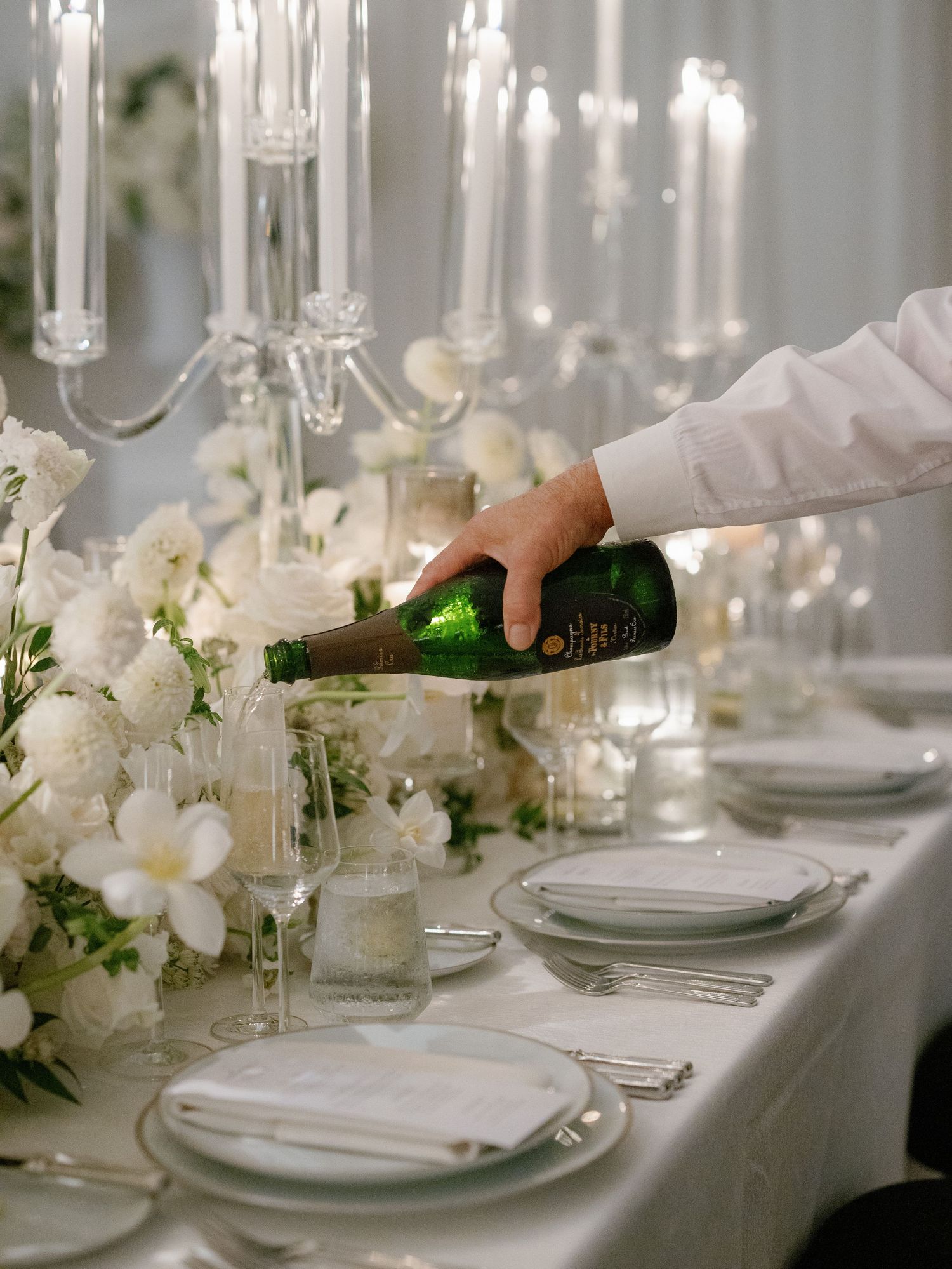 Server pouring champagne at elegant white wedding reception table with crystal candelabras.