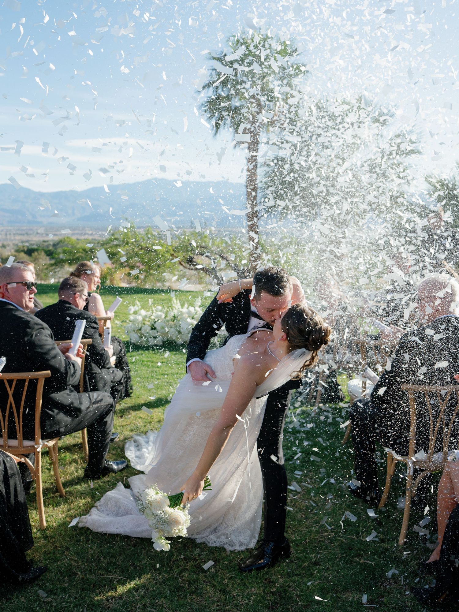 Groom dips bride for a kiss as confetti fills the air during outdoor wedding ceremony recessional.