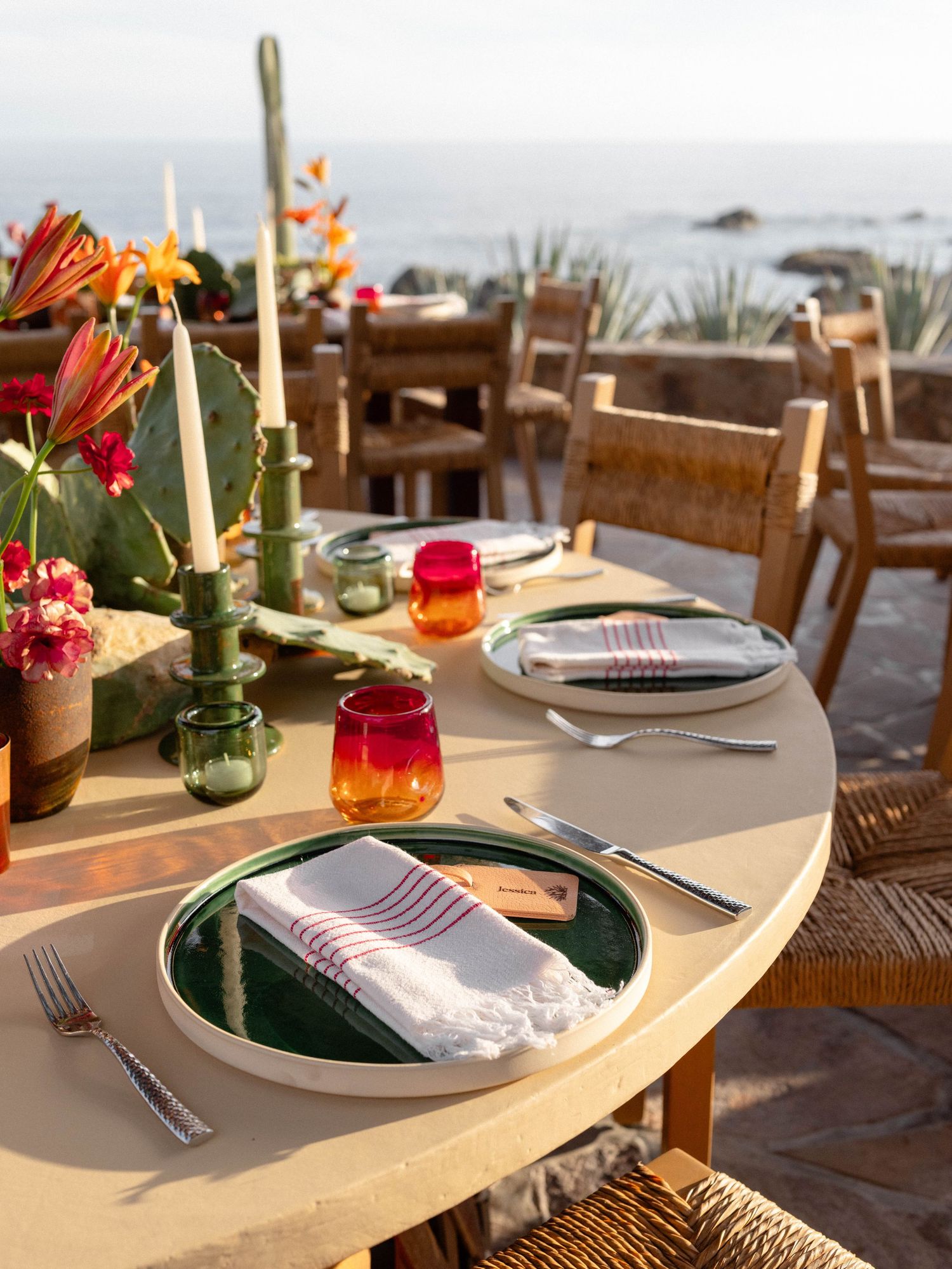 Elegant outdoor dining table set with green plates, striped napkins, candles, and colorful flowers overlooking the ocean.
