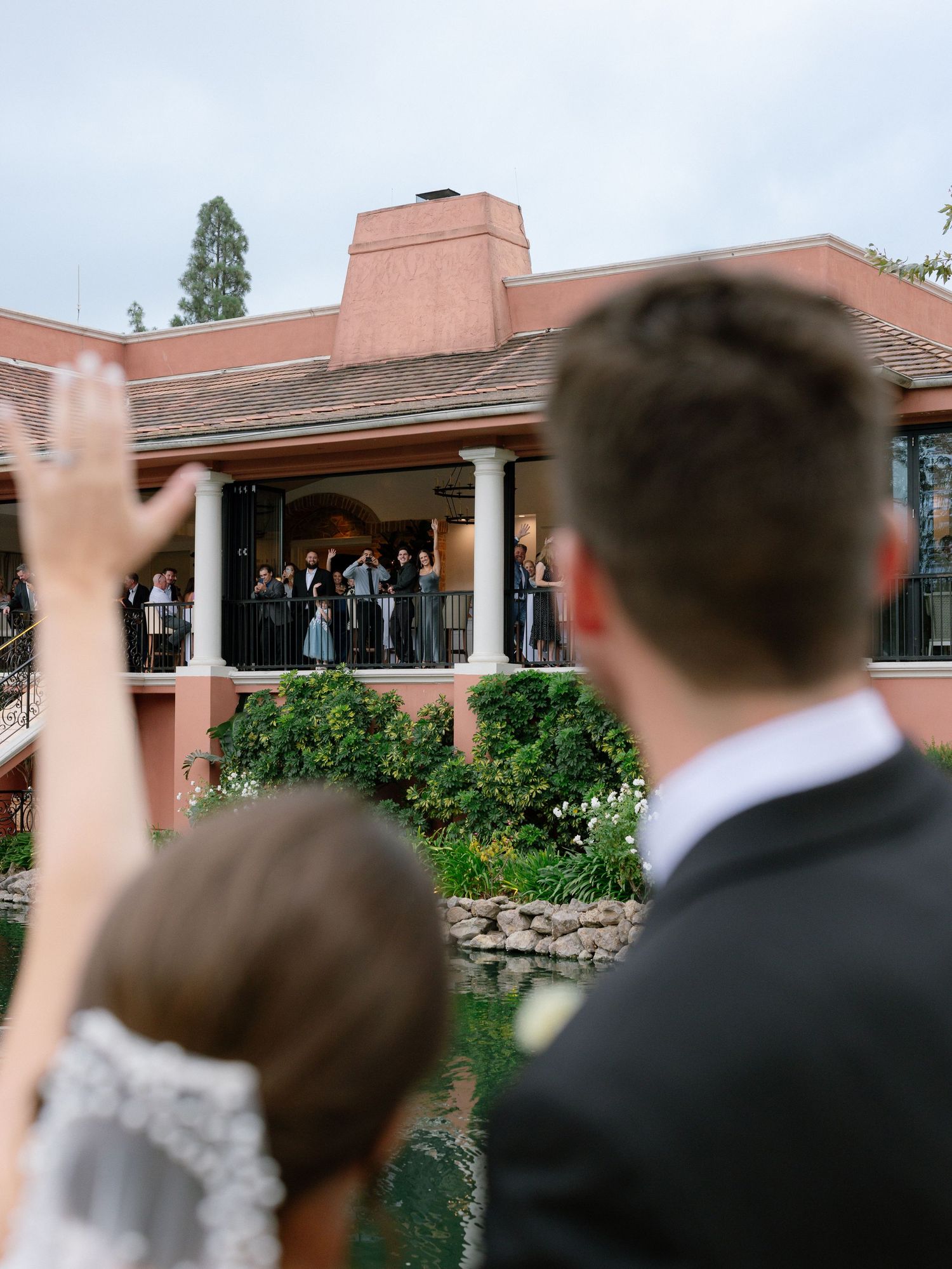 Bride and groom wave to wedding party gathered on balcony of elegant venue overlooking a pond.