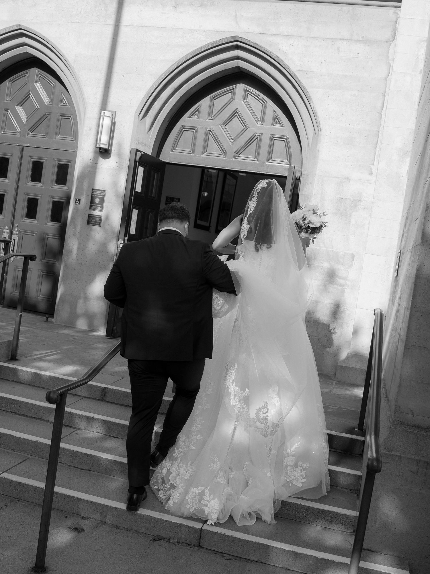 Bride and escort walk up stone steps entering church through ornate Gothic arched doors.