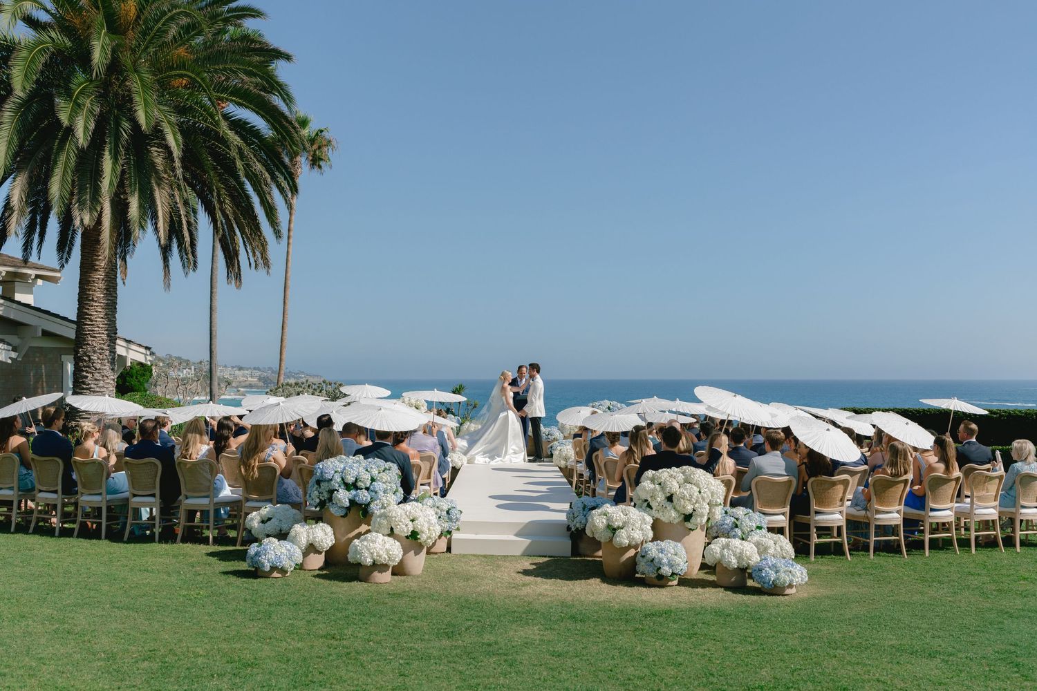Outdoor beachside wedding ceremony with guests seated under white umbrellas, blue hydrangeas, and a couple at the altar.