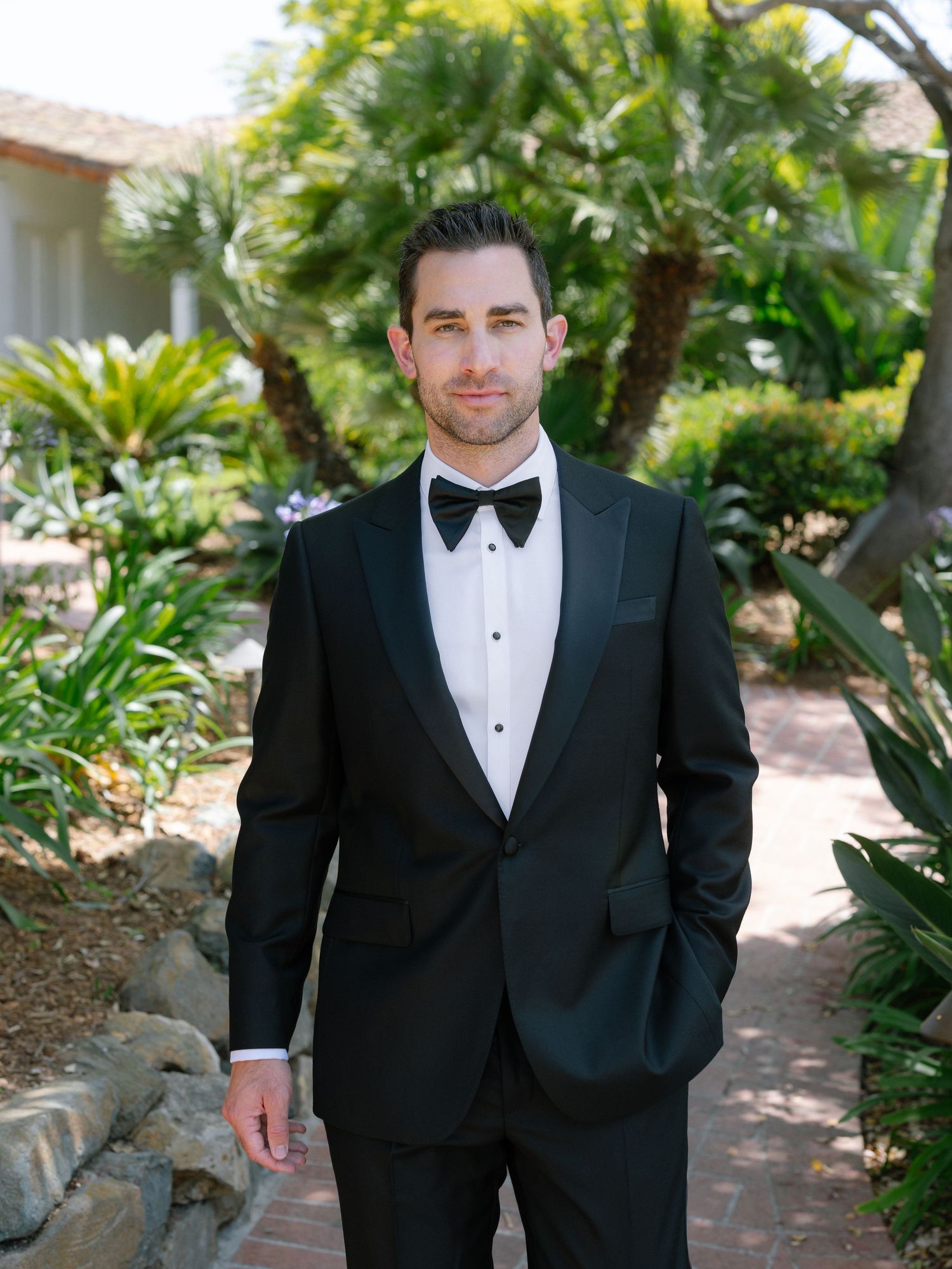Groom in classic black tuxedo with bow tie posing outdoors among tropical greenery.