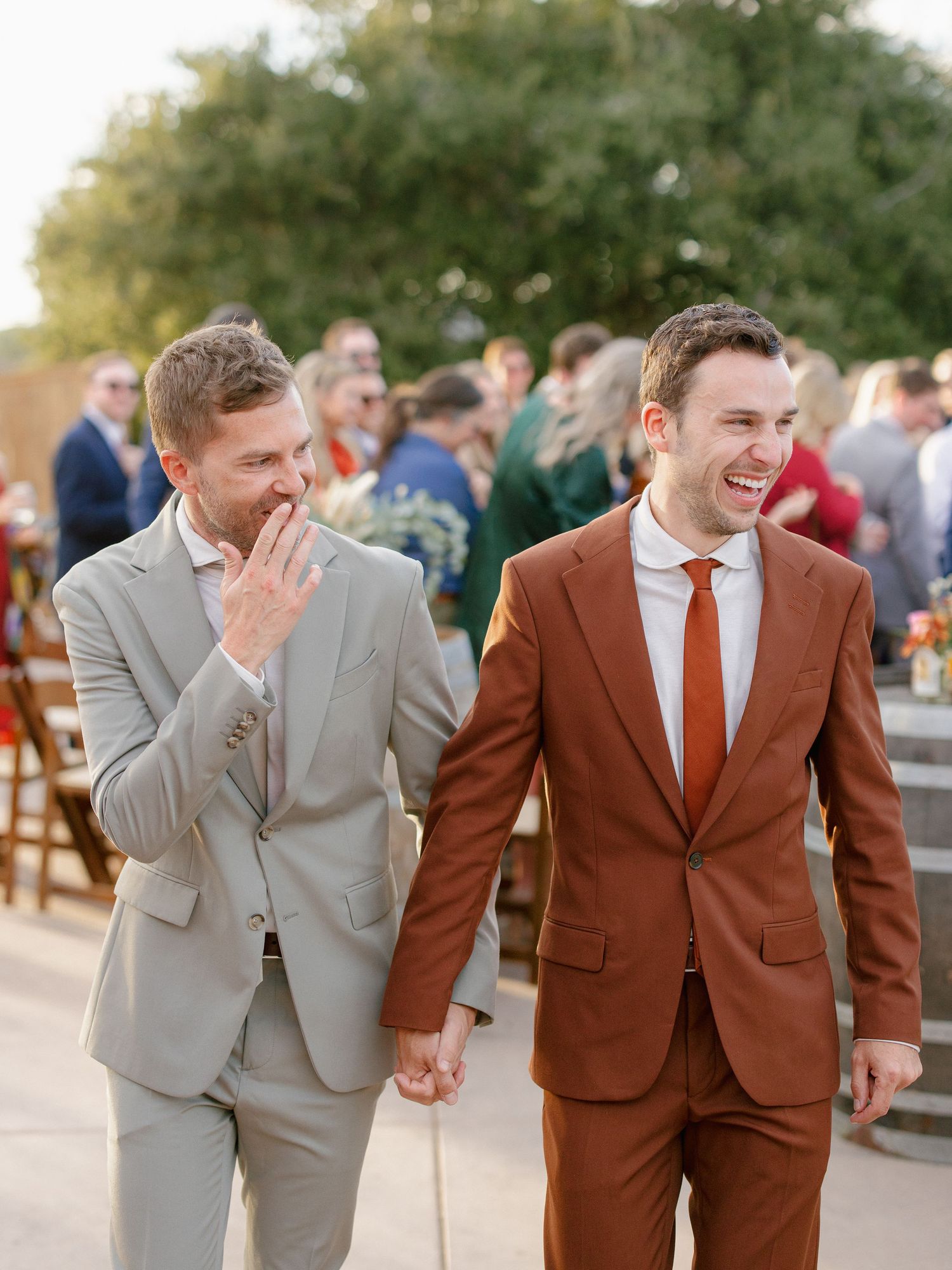 Two grooms in tan and rust suits laughing joyfully hand-in-hand at outdoor wedding celebration.