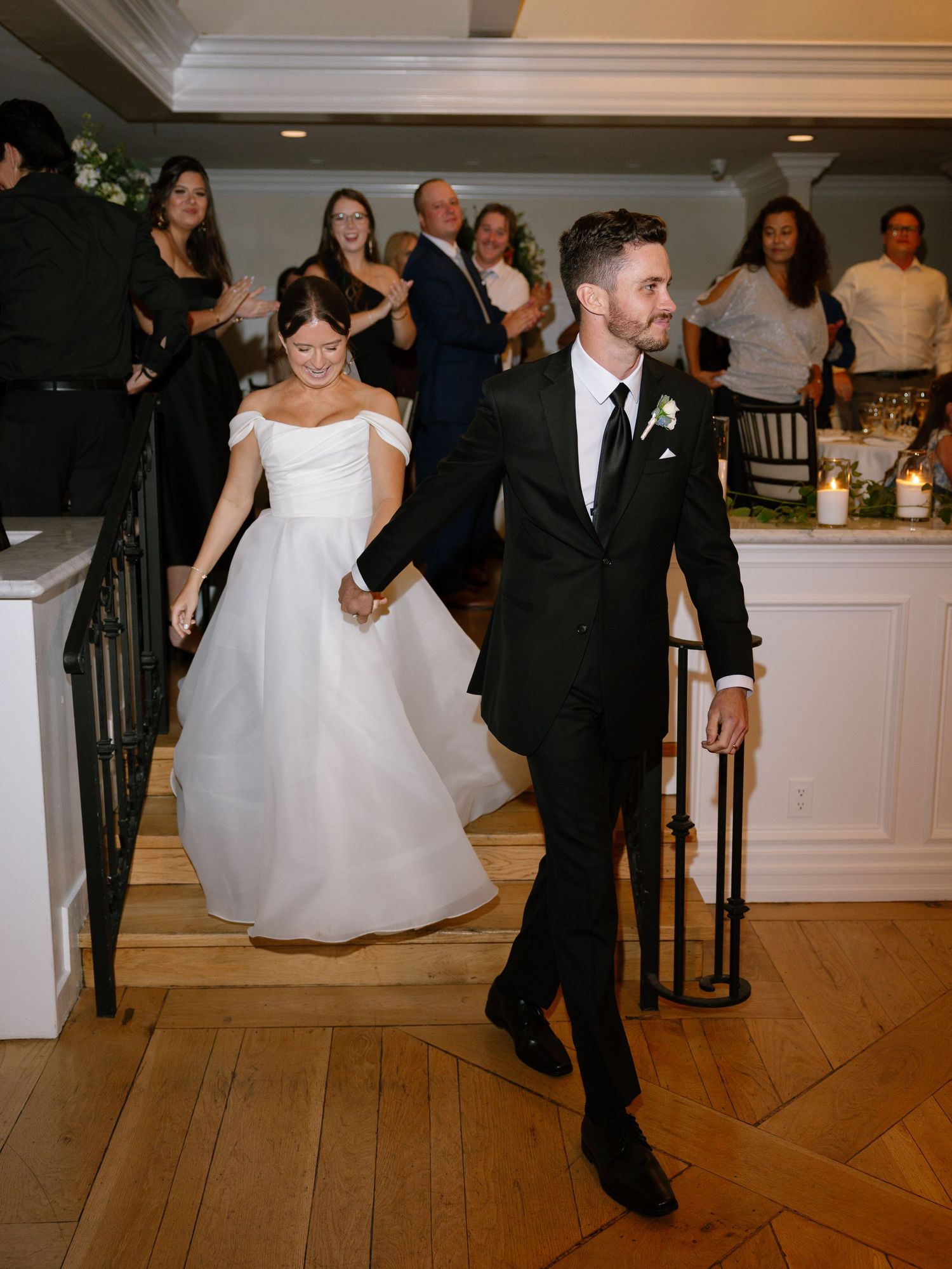Smiling bride and groom make grand entrance down staircase as guests cheer at wedding reception.