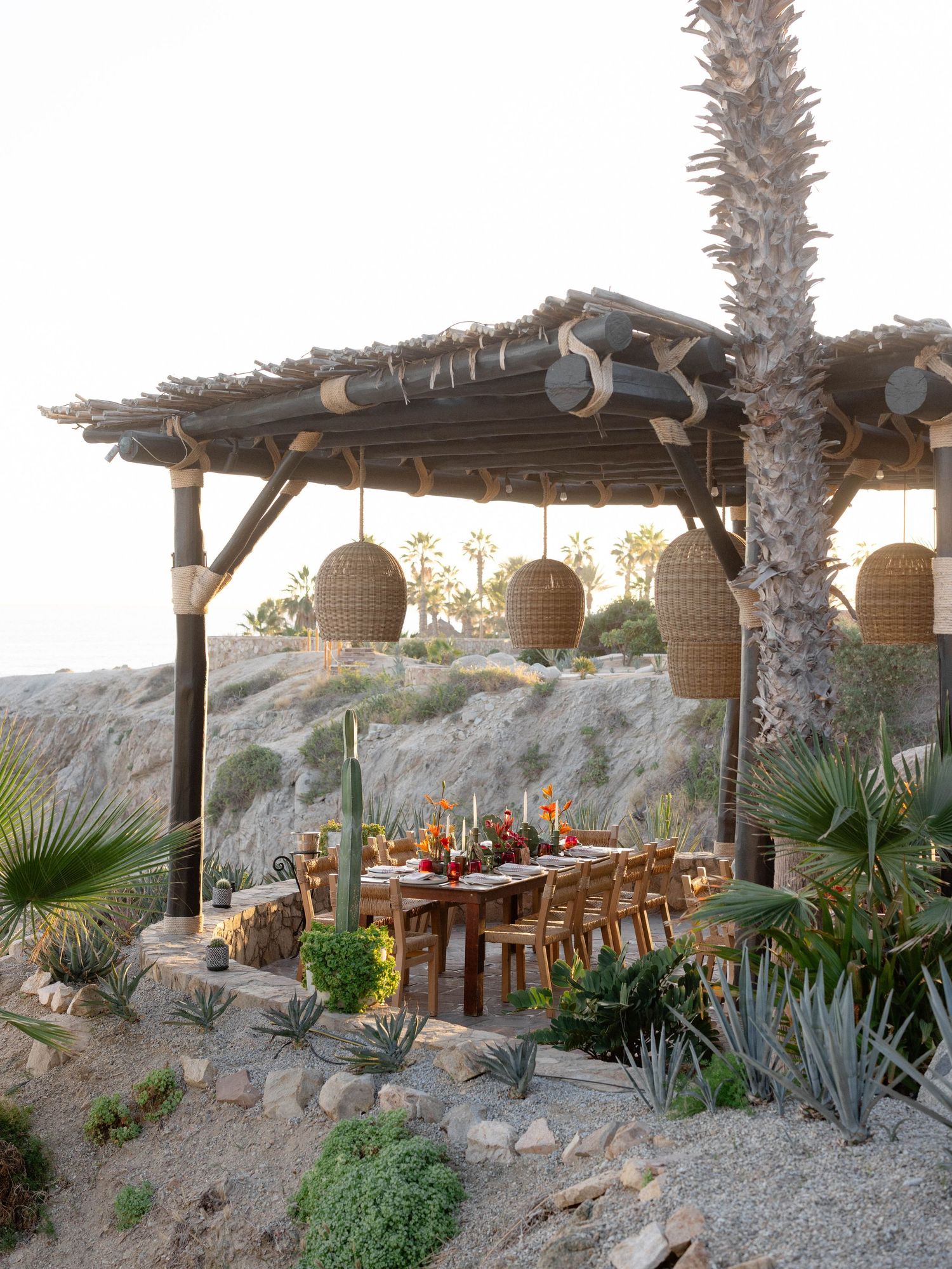 Rustic desert dining pergola with woven pendant lights, long wooden table, cacti, and agave plants at golden hour.