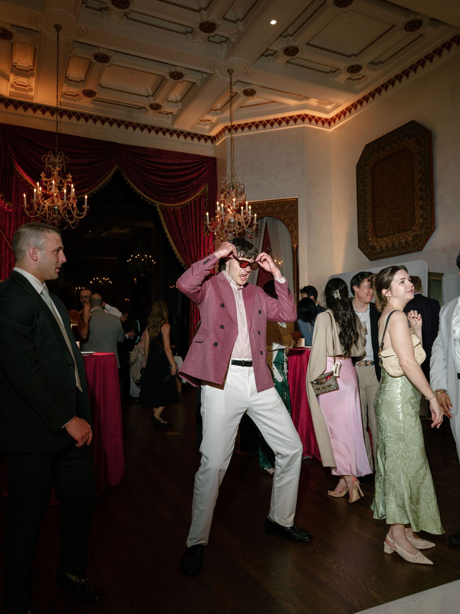 Guest in pink blazer dancing energetically at elegant wedding reception in ornate ballroom.