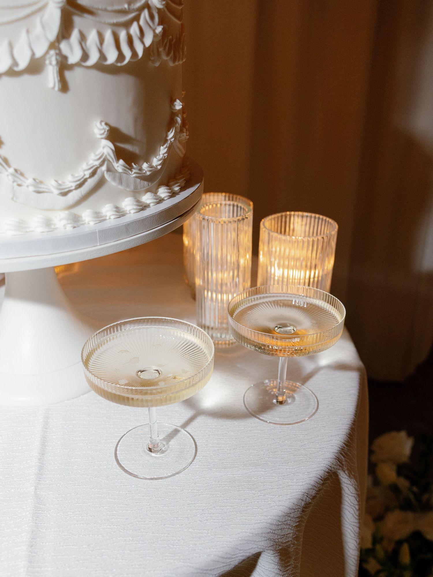 Wedding cake display with ribbed coupe glasses and gold votive candles on white linen.