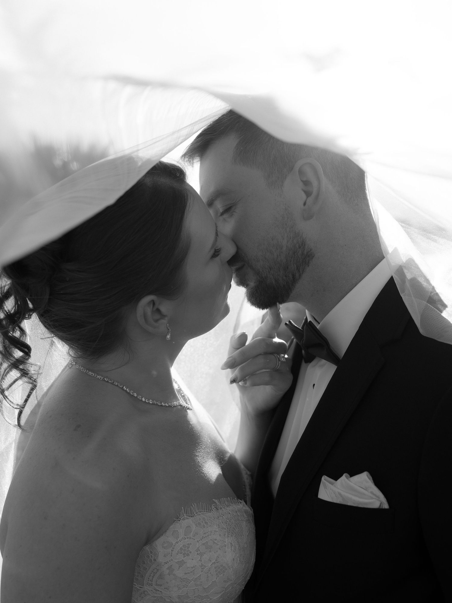 Black and white romantic photo of bride and groom nearly kissing under flowing wedding veil.