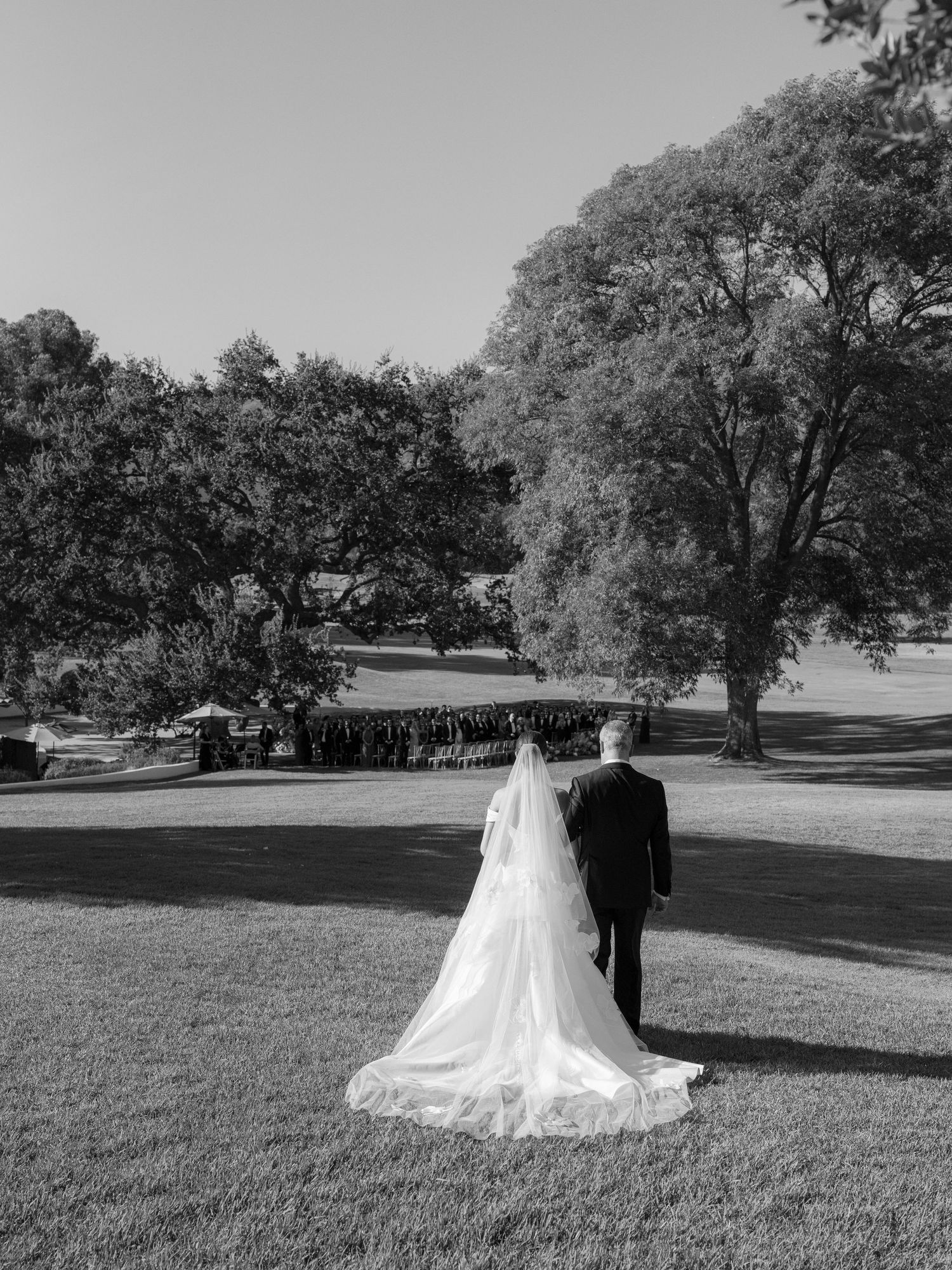 Black and white photo of bride in flowing veil and groom walking toward outdoor wedding ceremony.