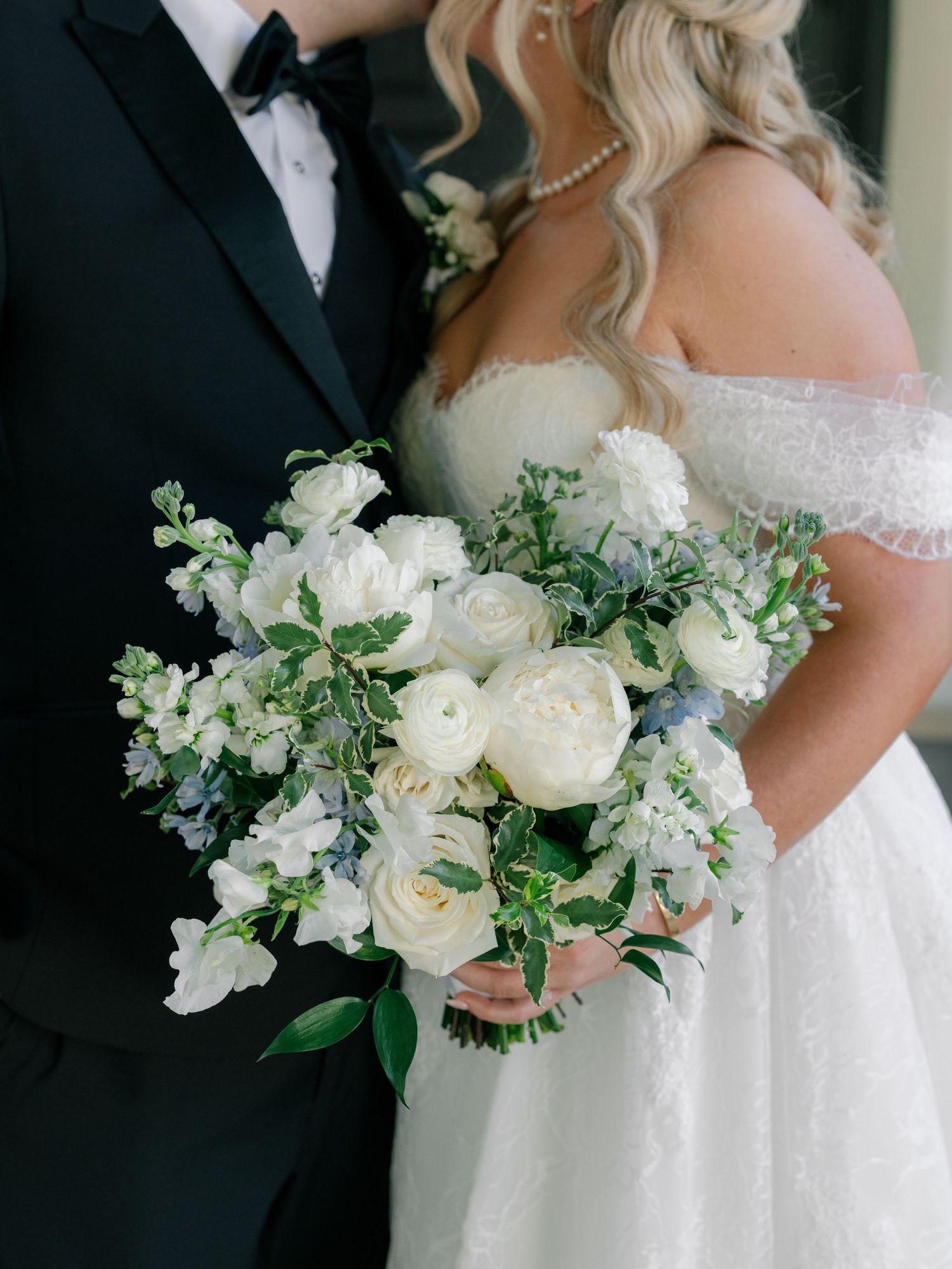 Bride holding a lush white peony and blue hydrangea bouquet with greenery beside groom in black tuxedo.