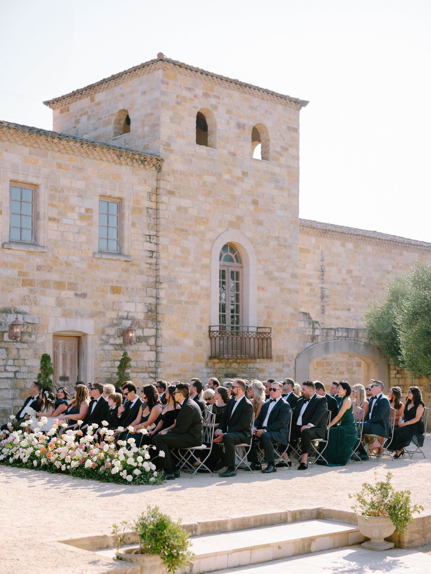 Wedding guests seated in rows before a rustic stone villa during an elegant outdoor ceremony with white floral decor.