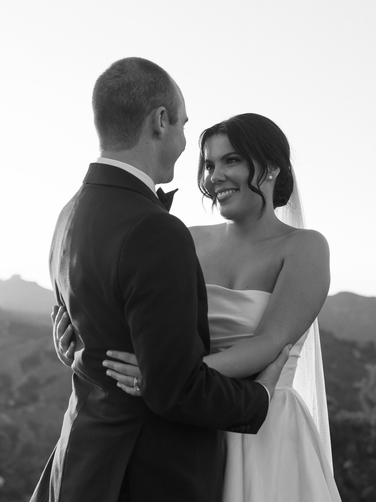 Bride and groom sharing a romantic black and white portrait embrace with mountain backdrop at sunset.