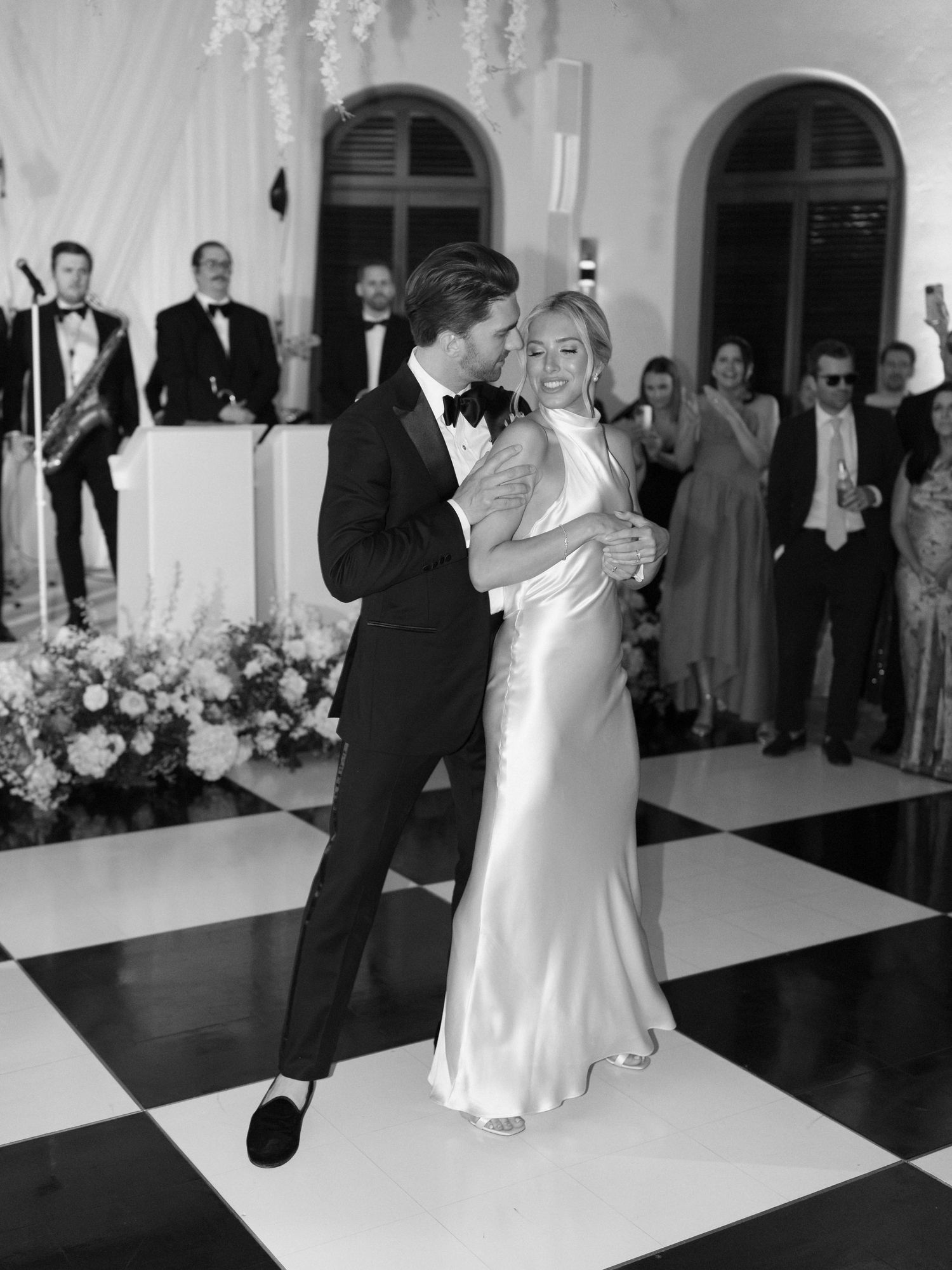 Couple shares first dance on black and white checkered floor, bride in silk gown, live band performing behind.