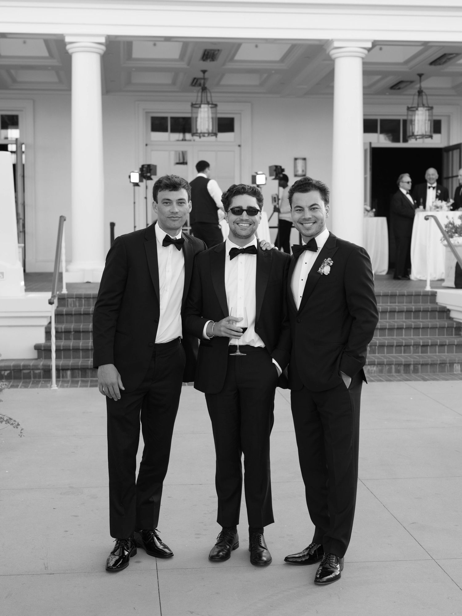 Three groomsmen in black tuxedos posing in front of a classic white columned venue exterior.