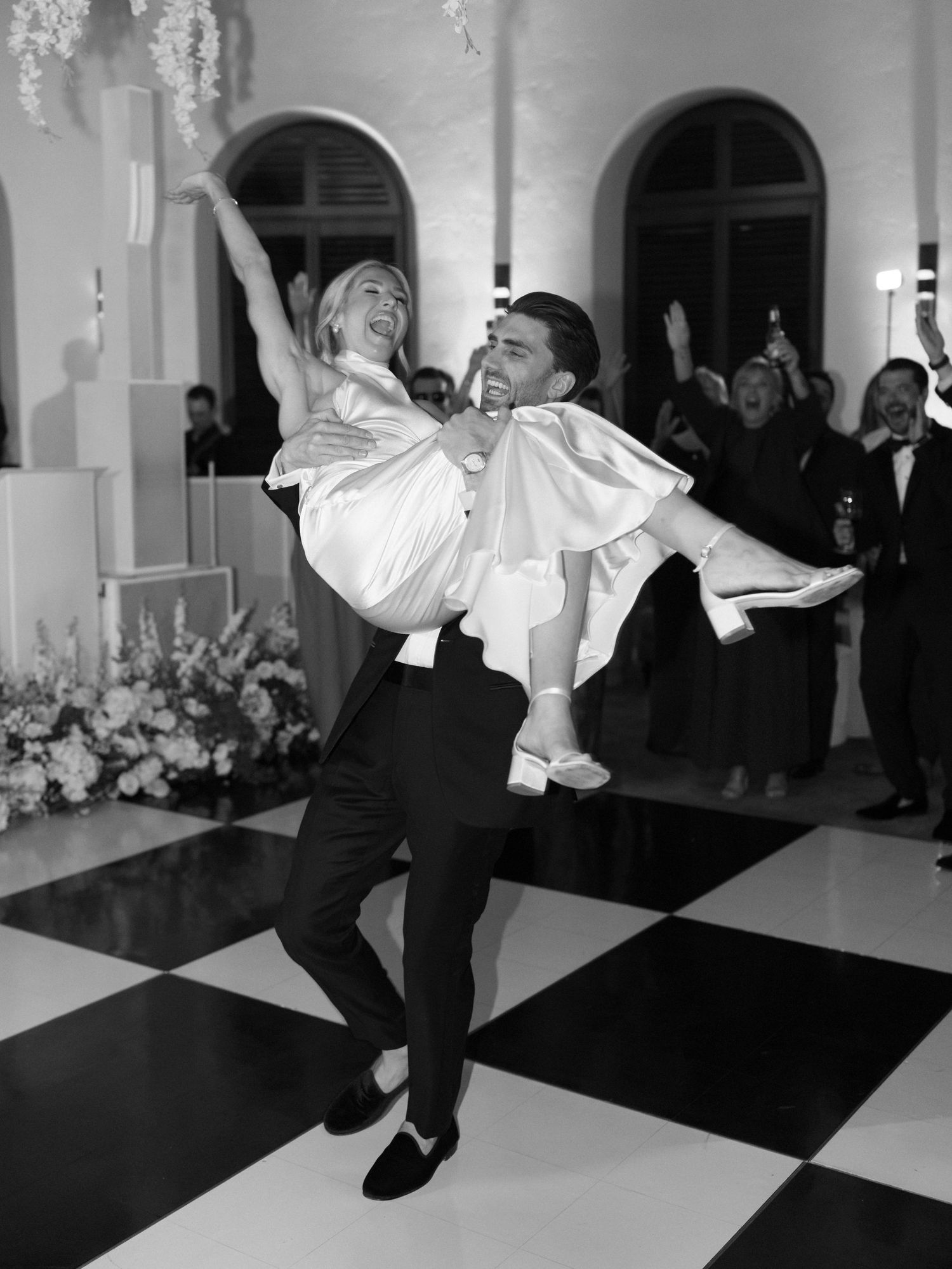 Black and white photo of groom joyfully lifting bride on a checkered dance floor during wedding first dance.