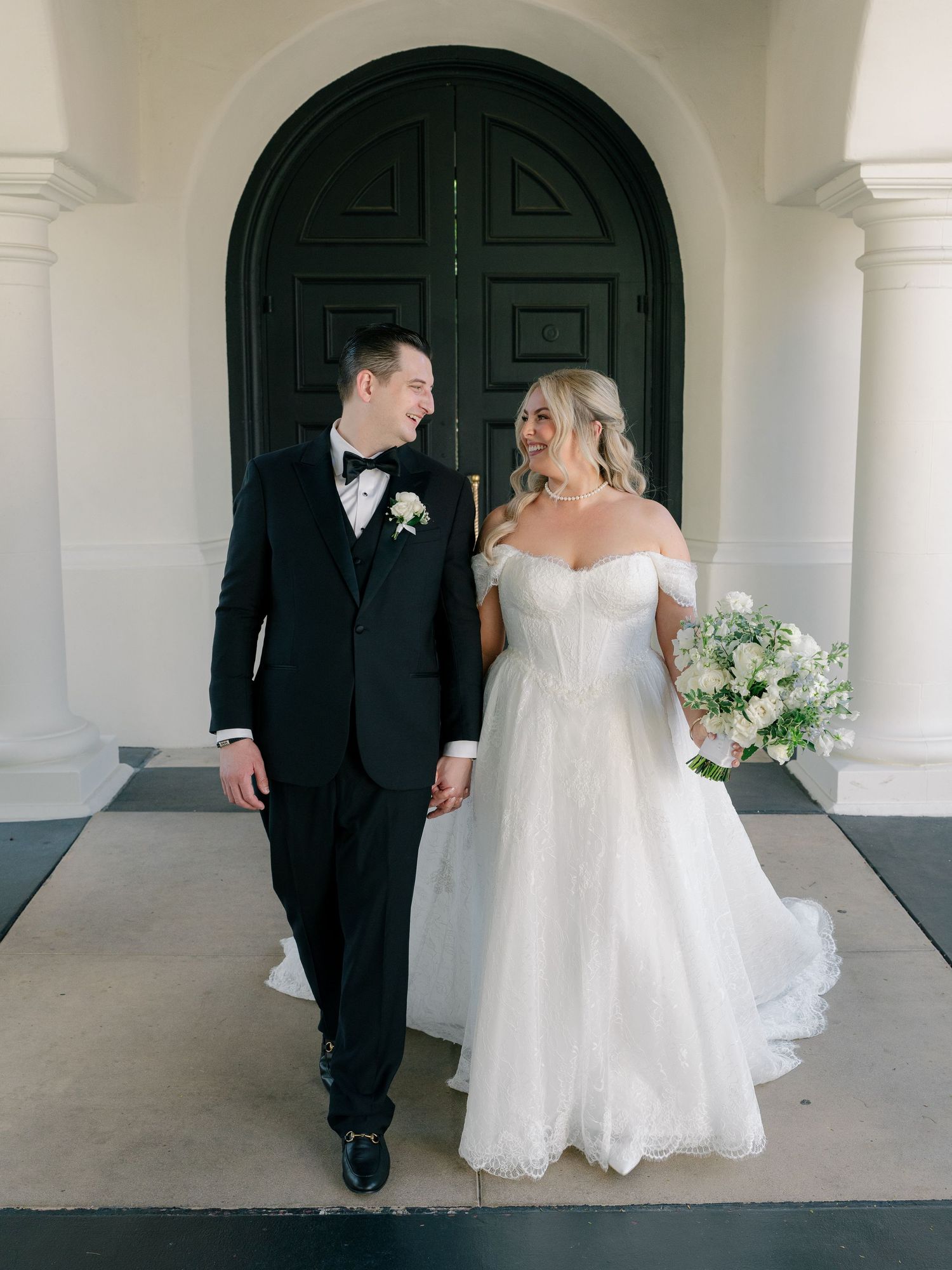 Bride in off-shoulder lace gown and groom in black tuxedo smiling at each other before dark arched doors.